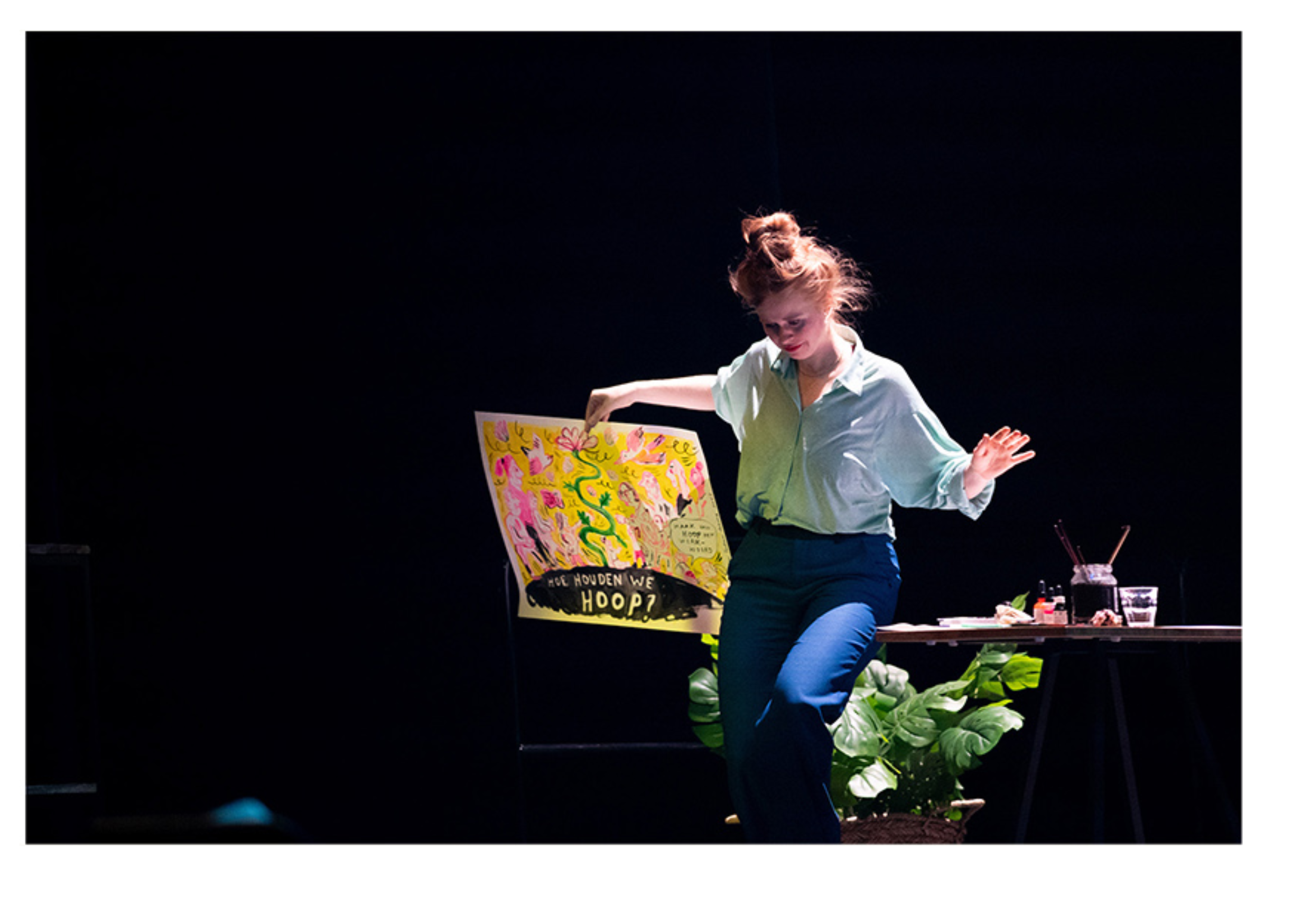 A young woman with red hair styled in a messy bun, wearing a light-colored shirt and dark pants, performing on stage with a colorful, illustrated poster and art supplies on a table beside her.