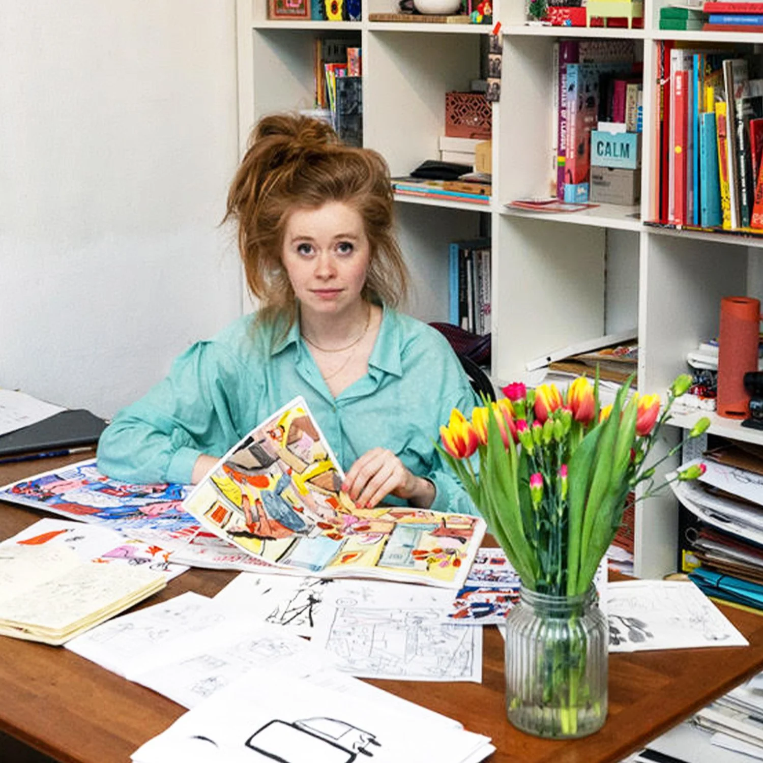 A woman with messy red hair sitting at a cluttered desk filled with papers, comics, and sketches, in front of white shelves filled with books and colorful objects, holding a comic book in her hand, with a vase of pink and yellow tulips on the desk.