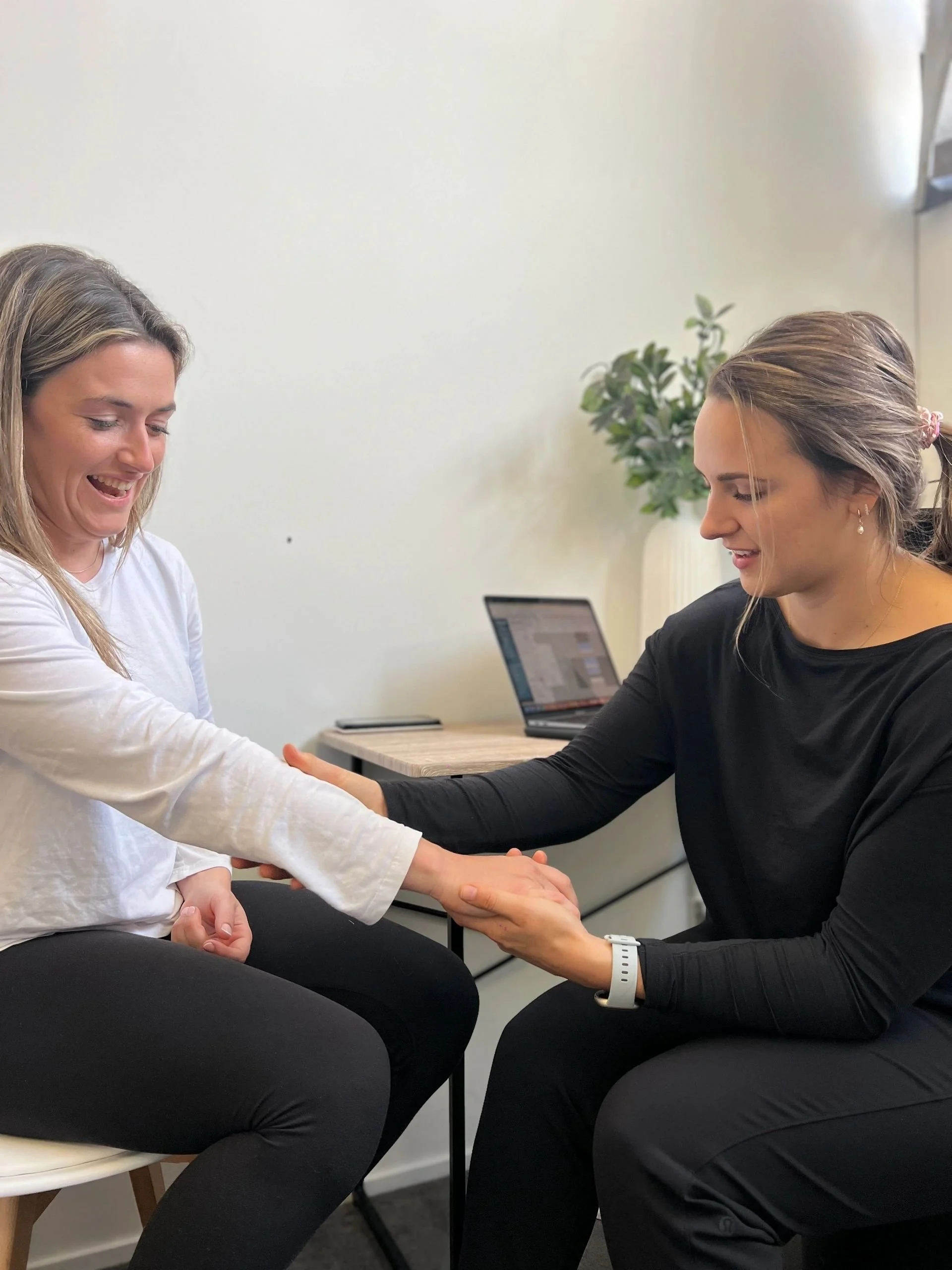 A woman in black sitting and holding hands with a woman in white who is smiling, in an office with a laptop and a potted plant in the background.