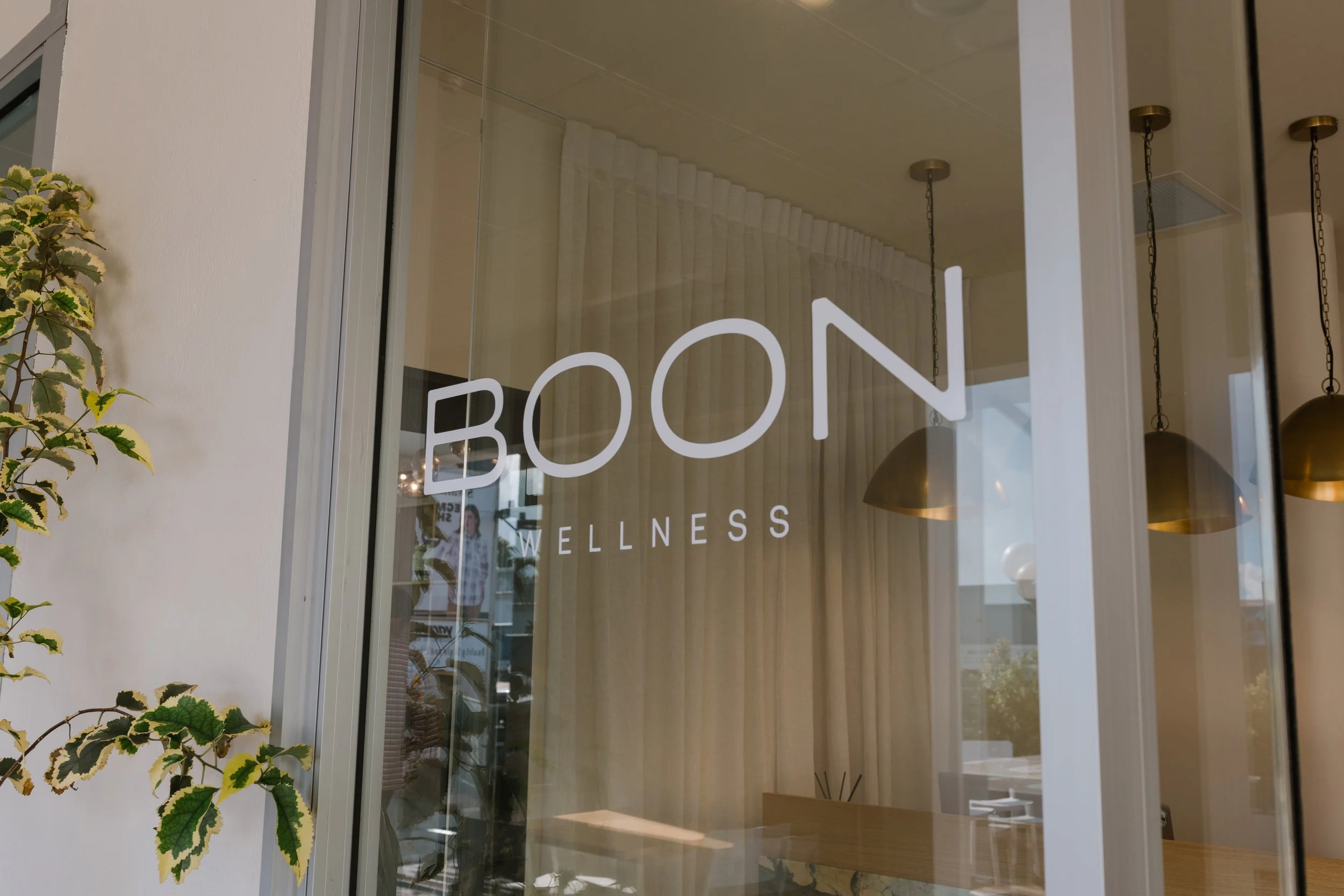 View through a glass door with the words 'BOON WELLNESS' written on it, showing the shop's interior with hanging pendant lights and beige curtains.