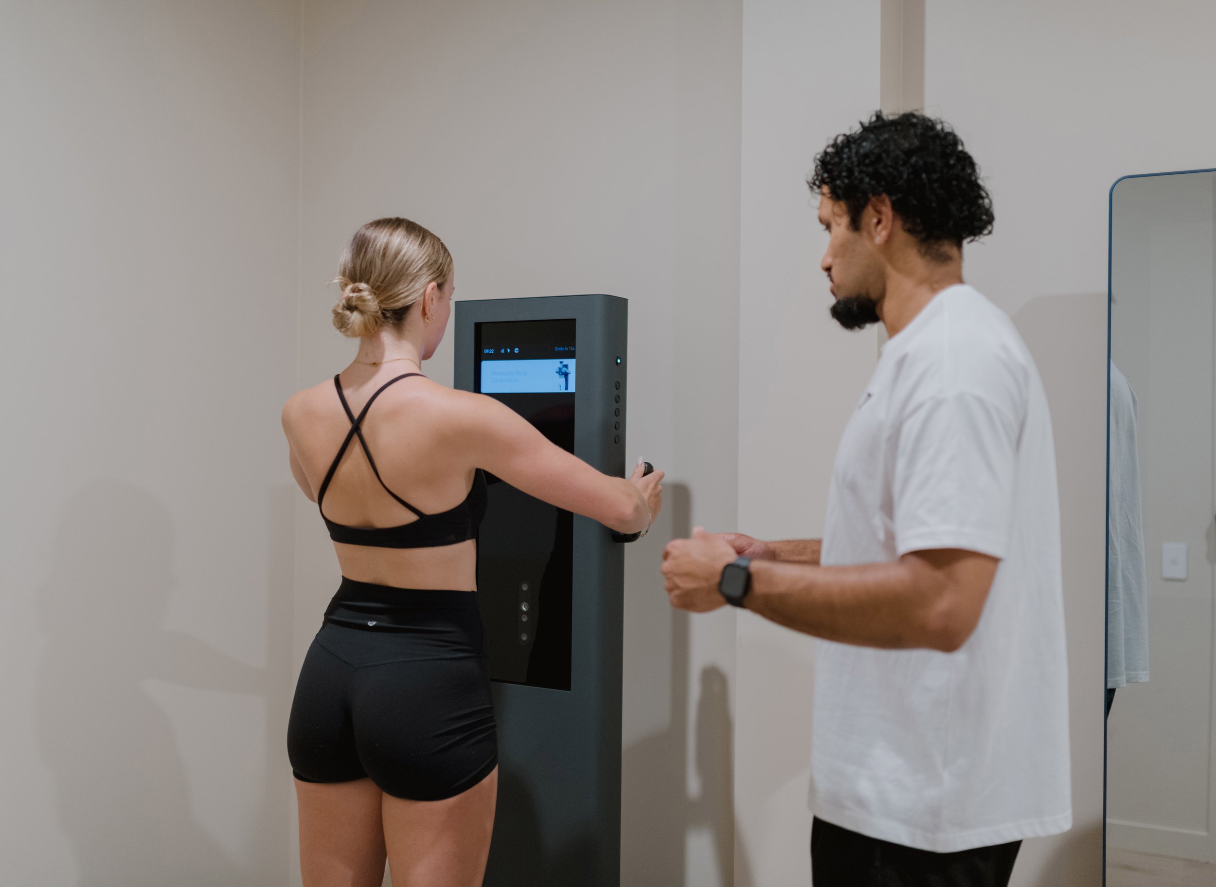 A woman in black sportswear uses a modern fitness assessment machine under supervision of a male healthcare professional in a clinical setting.