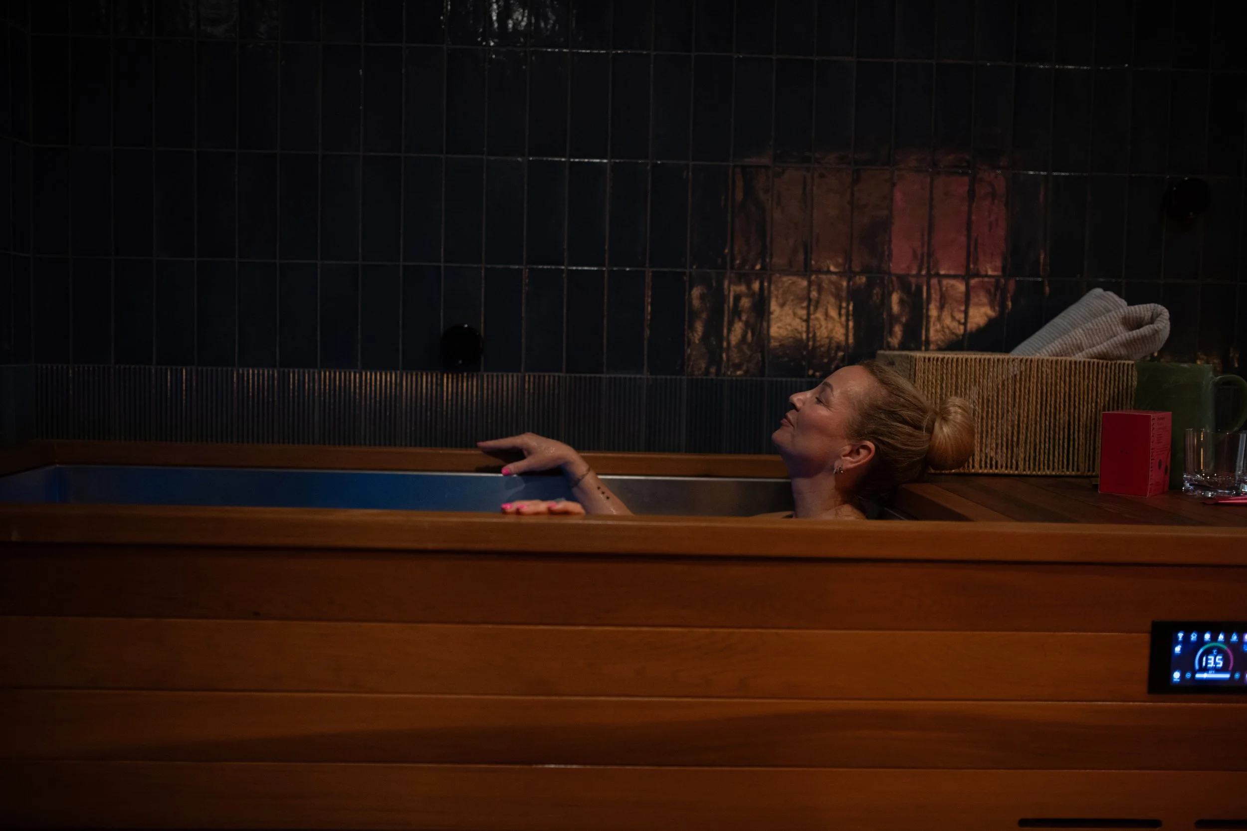 Woman relaxing in a wooden ice tub with closed eyes in a dimly lit room with dark tiled walls.