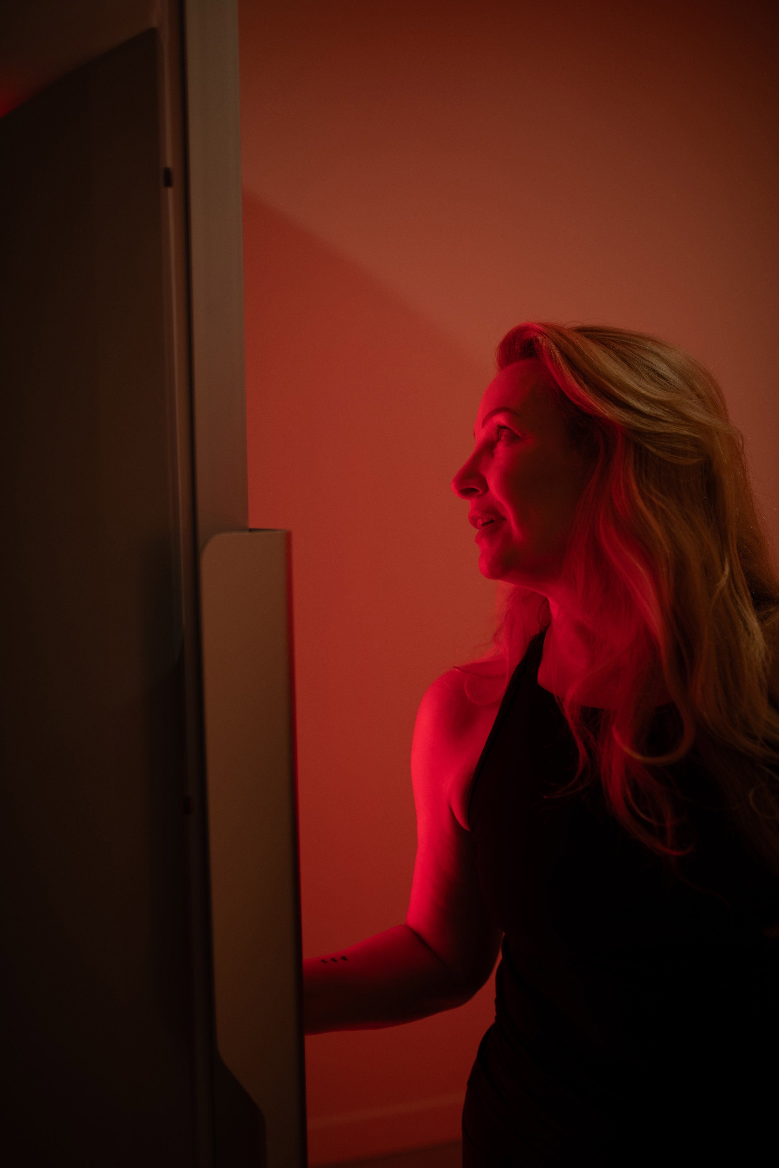A woman with wavy blonde hair looking into a red light therapy booth.