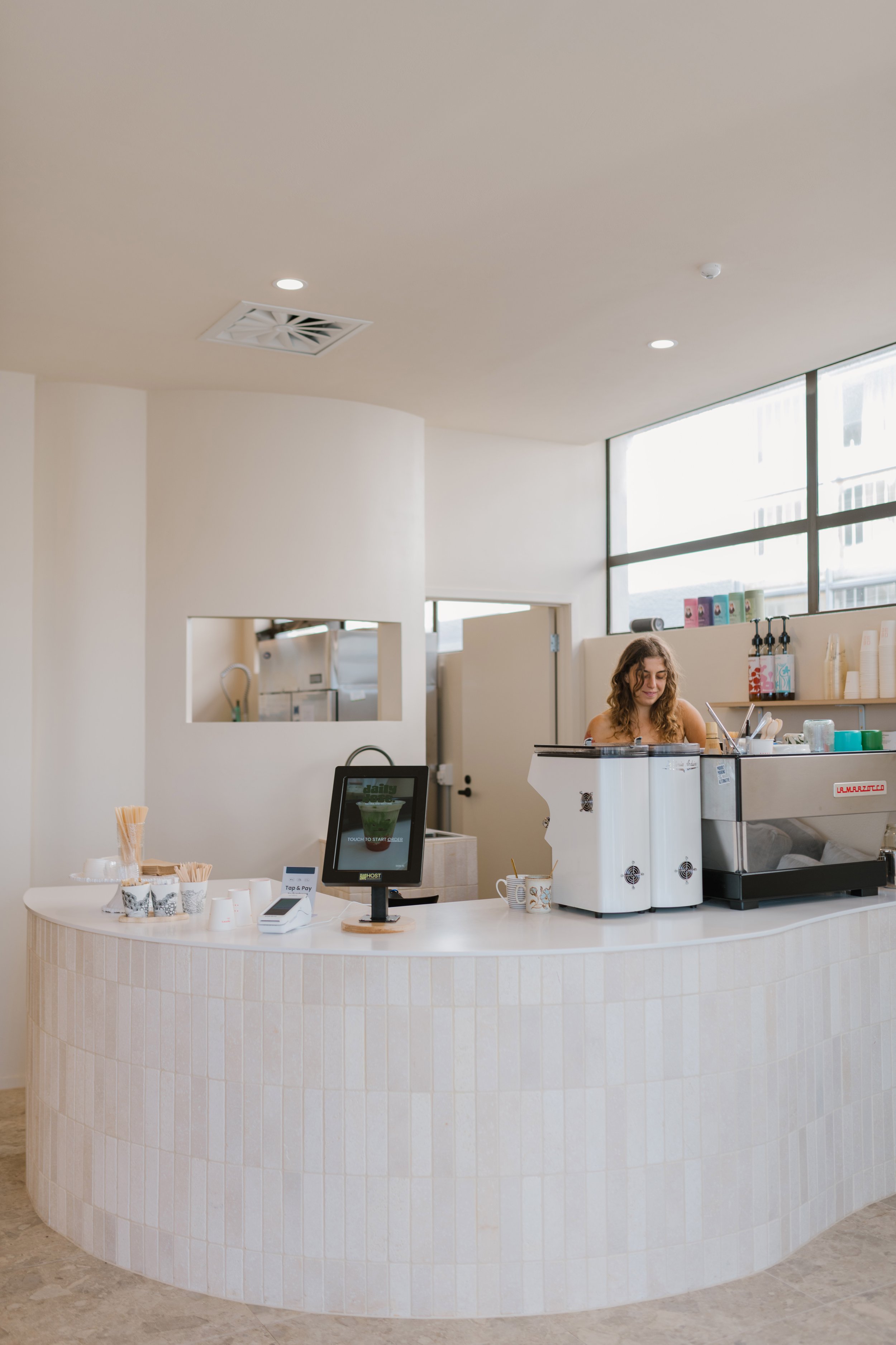 A woman standing behind a modern, curved coffee counter in a well-lit cafe, with a coffee machine and cups in front of her.