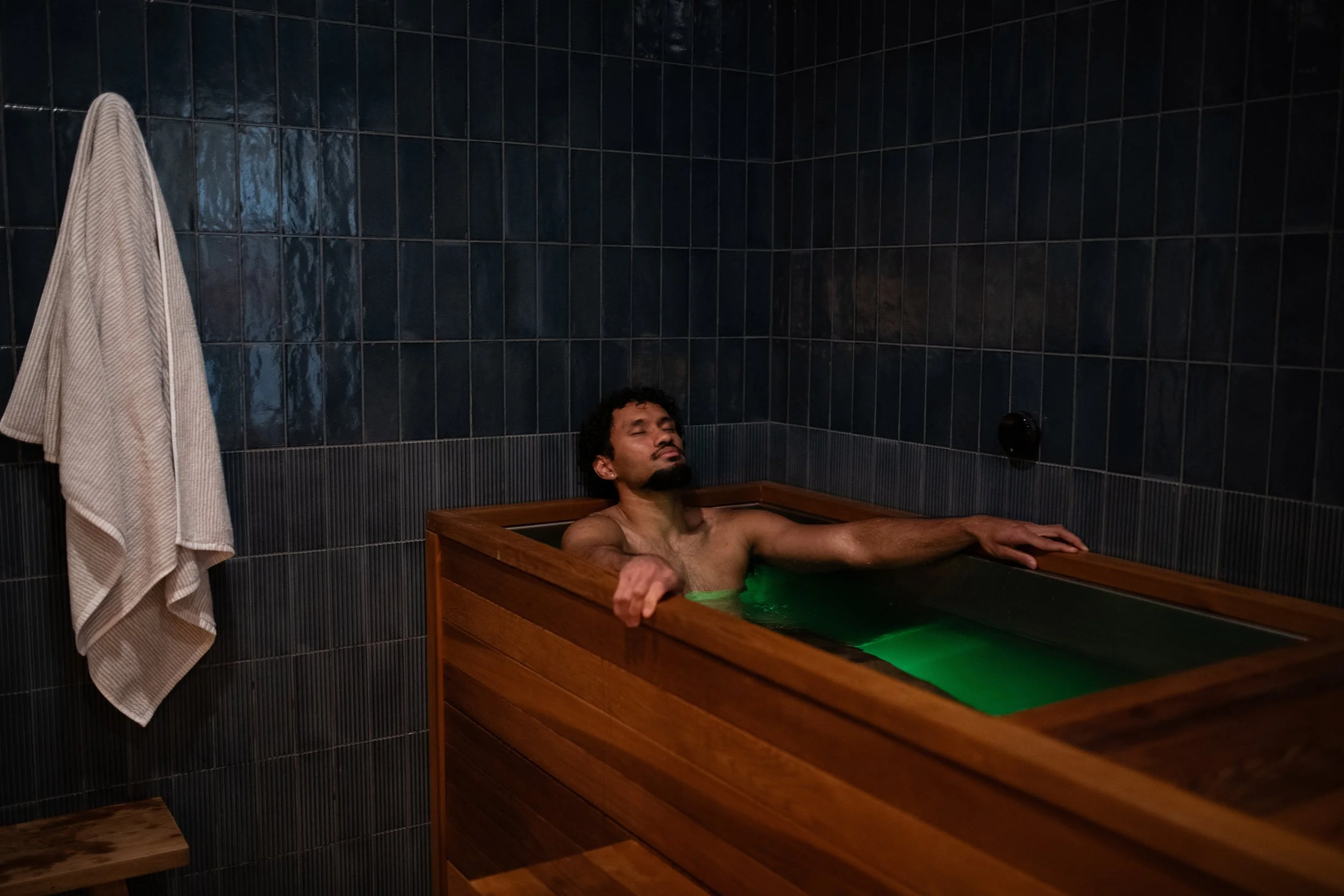 A man relaxing in a wooden soaking bath with green-colored water in a dark tiled room.