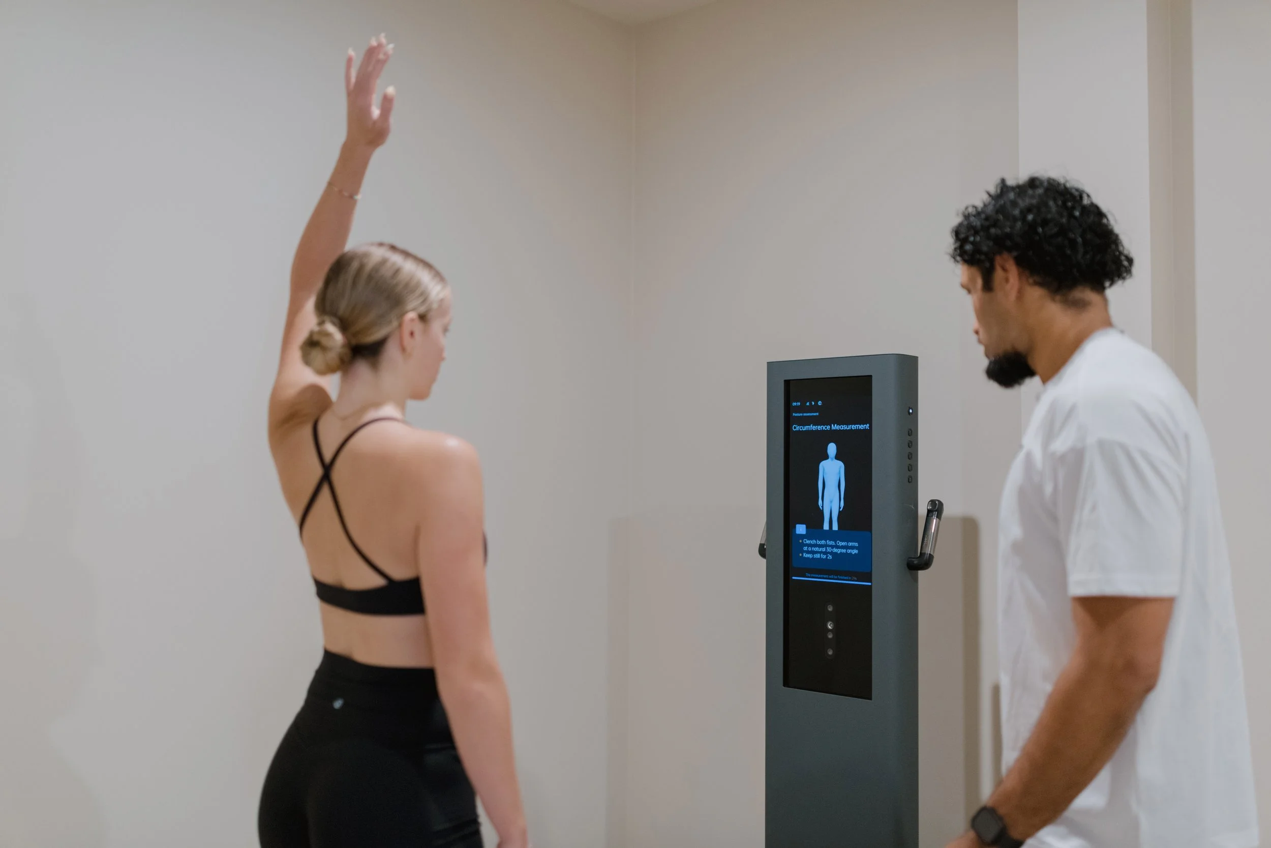A woman participating in a body measurement exam while a technician observes. The woman has her arm raised, and the technician is looking at a digital screen displaying a human figure and measurement instructions.