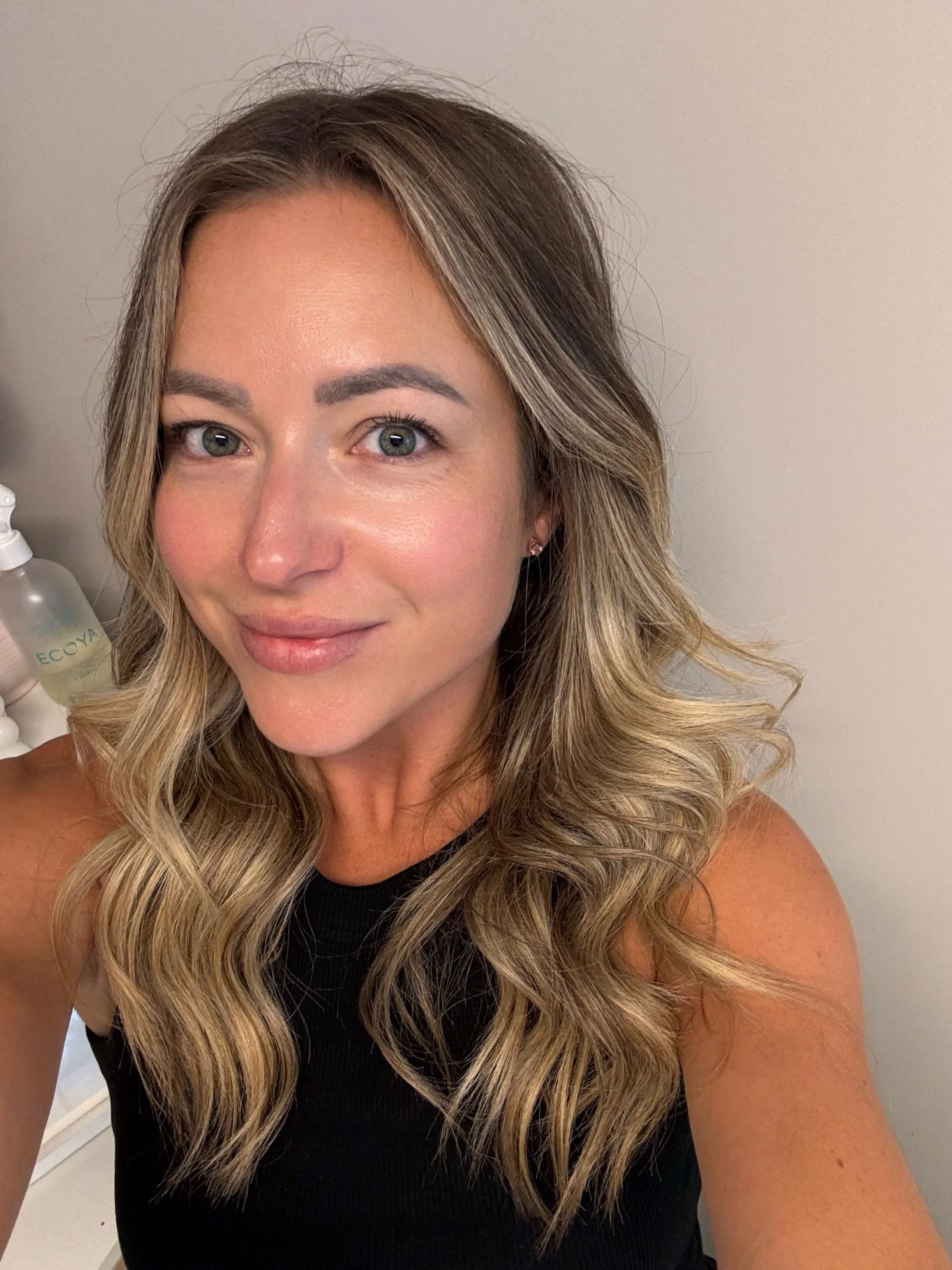 A woman with long, wavy blonde hair and bright blue eyes smiling at the camera. She is wearing a black sleeveless top and has small earrings. The background is plain and beige.