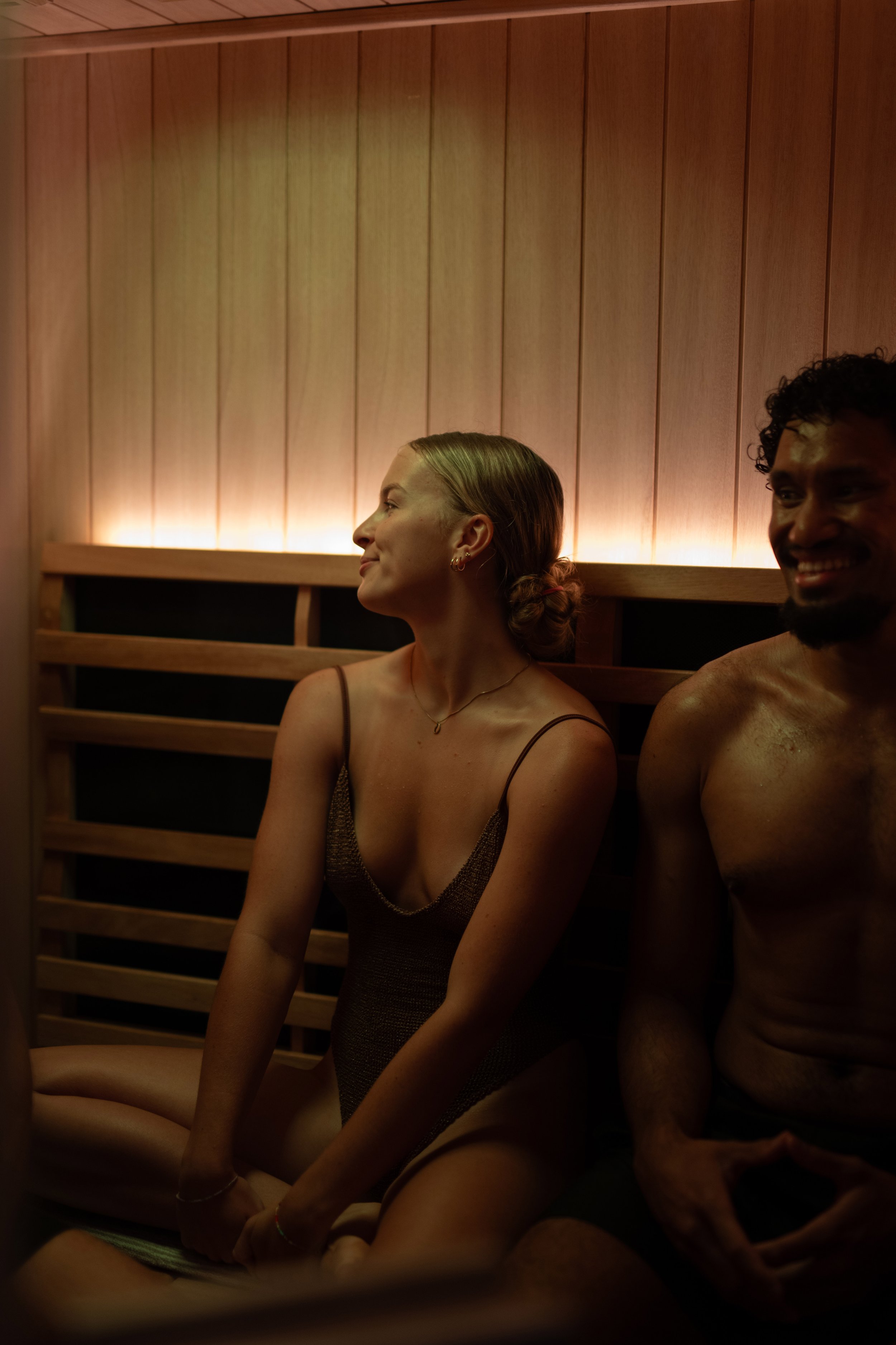 Two smiling people sitting in a sauna with wooden walls, one woman with blonde hair and a swimsuit, and one shirtless man with curly hair.