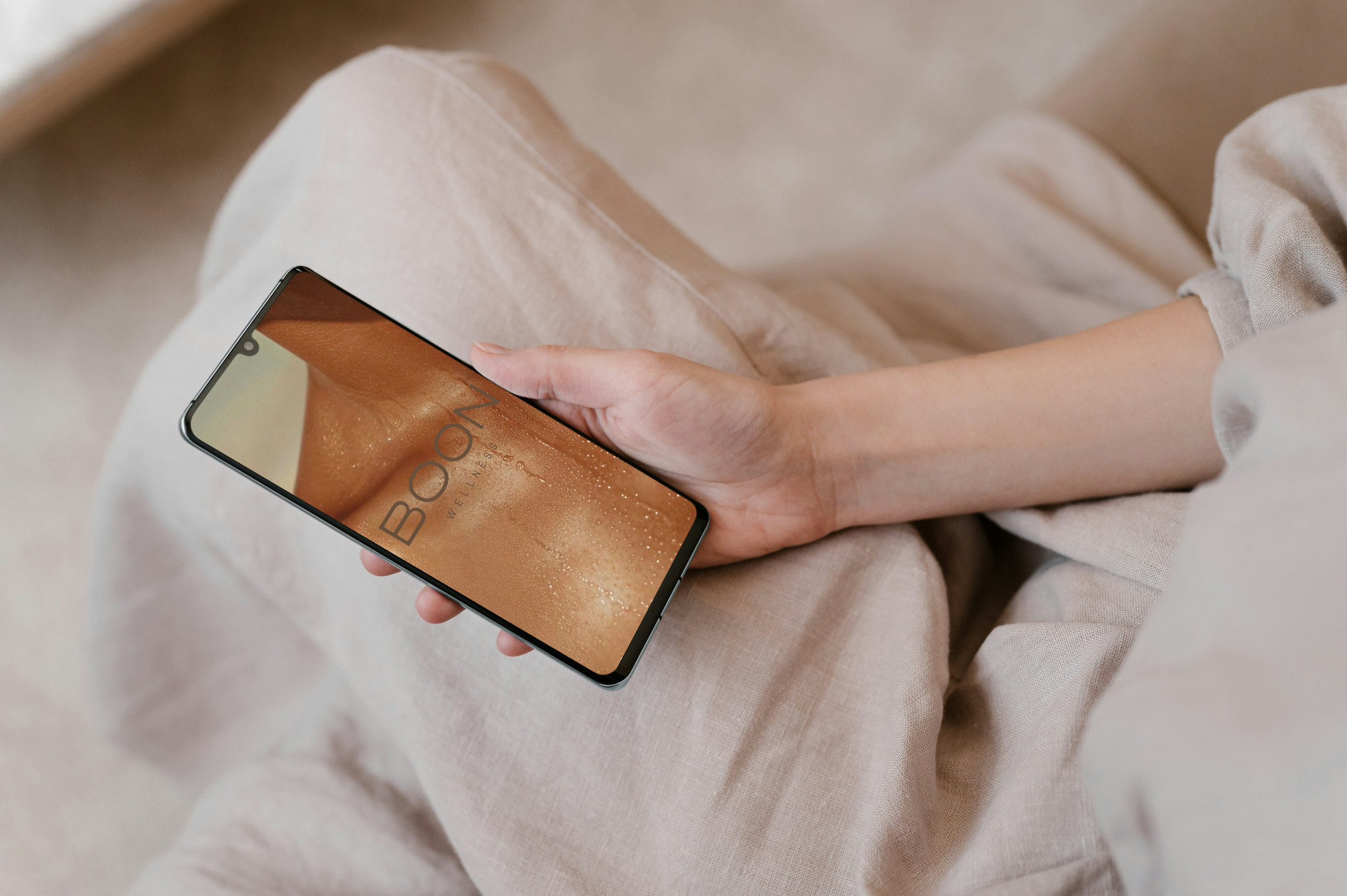A person sitting on a beige fabric chair or sofa, holding a smartphone with the BOON Wellness app on the screen.