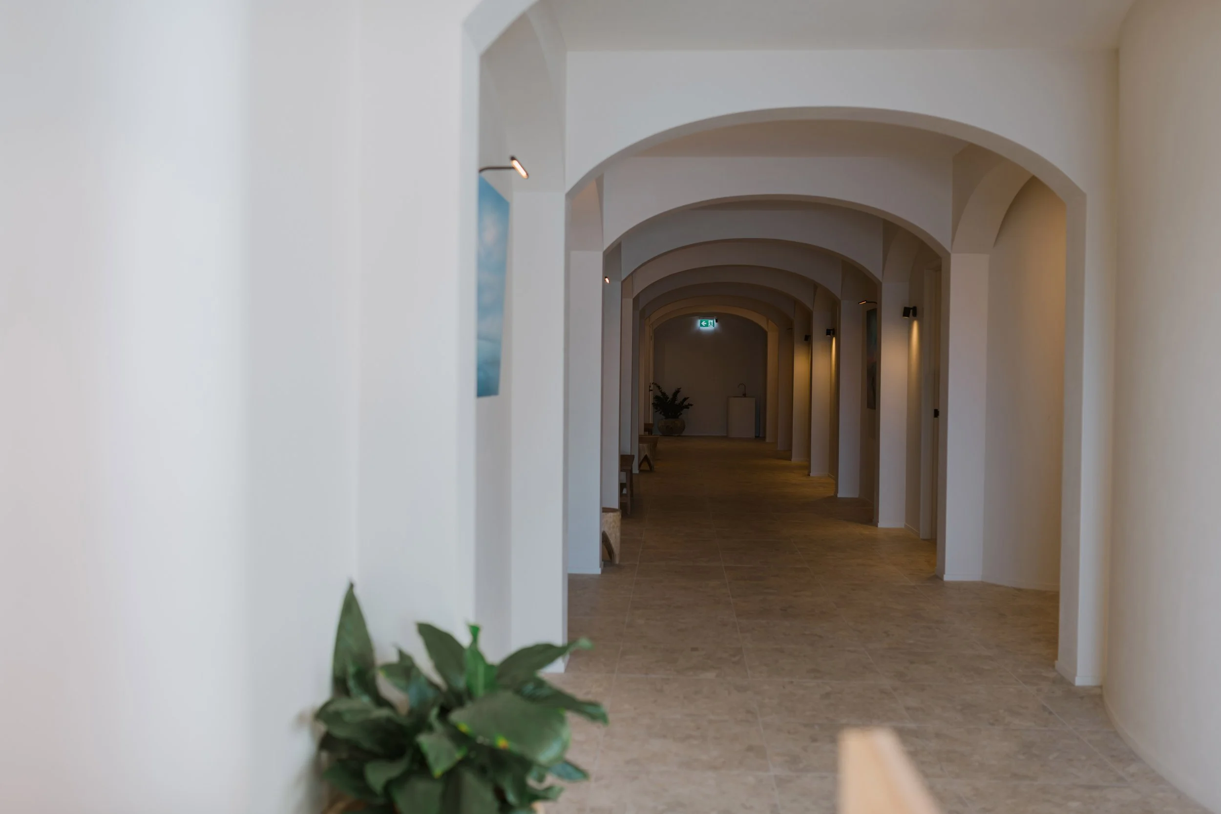 Long hallway with beige tile floor, white arched ceiling, and soft lighting, with a potted plant at the far end and framed pictures on the walls.