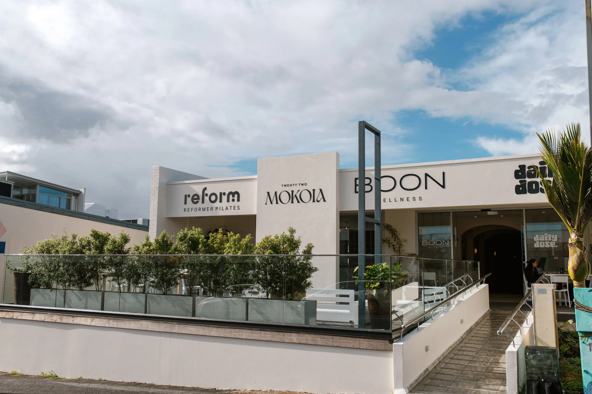 Modern building with signage for 'reform', 'MOKUA', and 'BOON', featuring a walkway with a railing, outdoor plants, and a person sitting at a table inside; cloudy sky above.