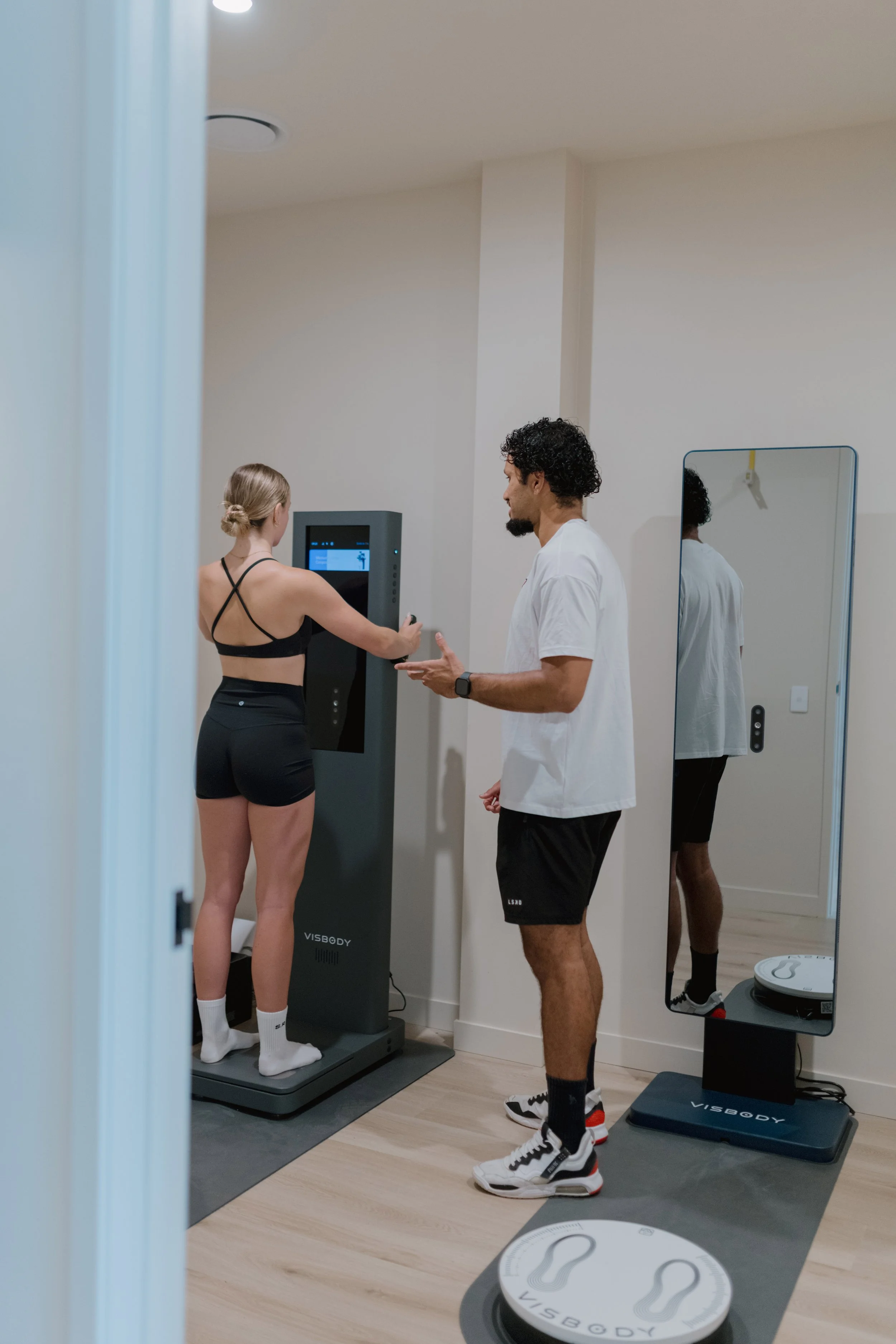 A woman in athletic wear standing on a body composition scale while a male technician monitors her data in a fitness assessment room.