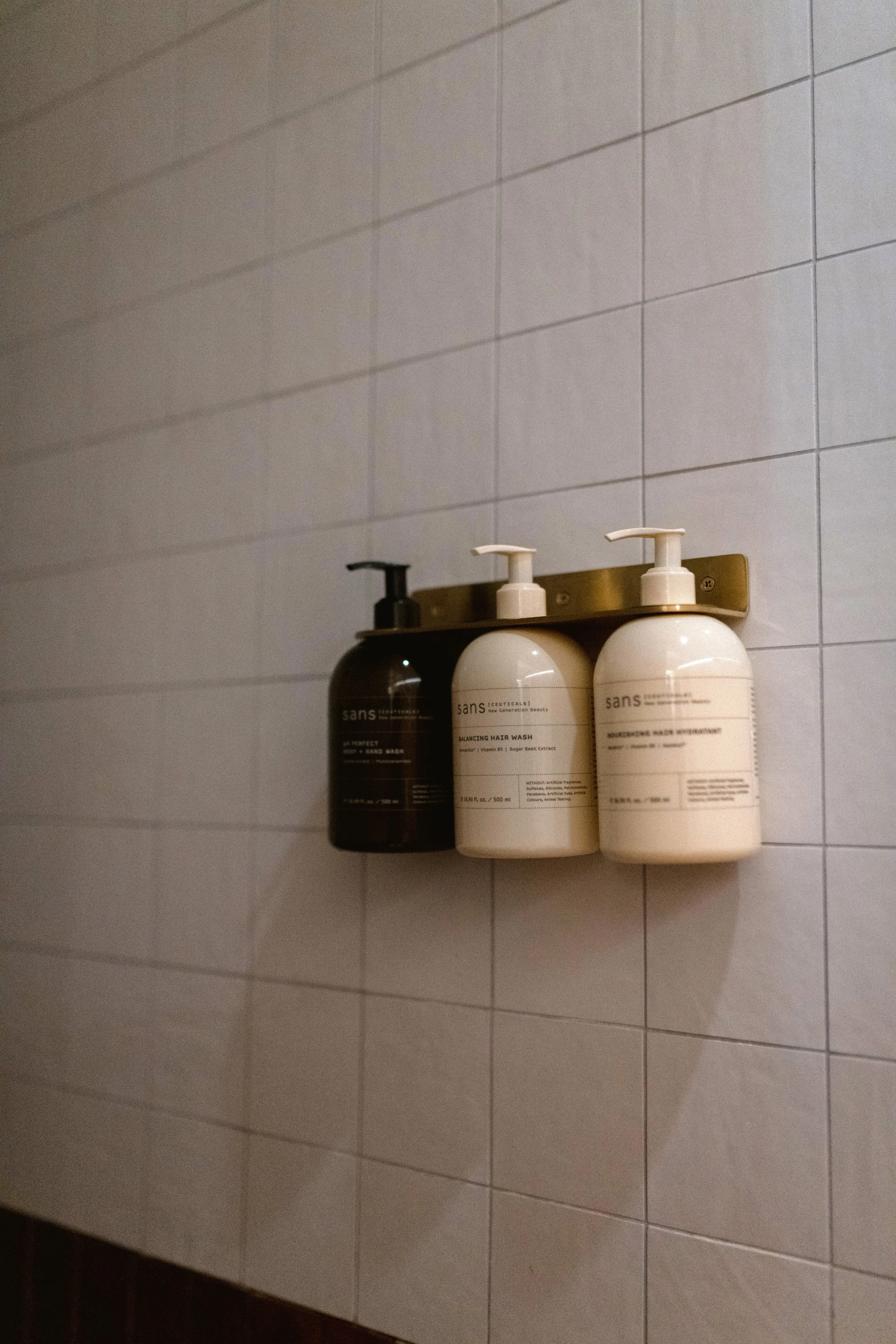 Three bottles of toiletries on a metal shelf mounted on a tiled bathroom wall, with the bottles having pump dispensers and labels.