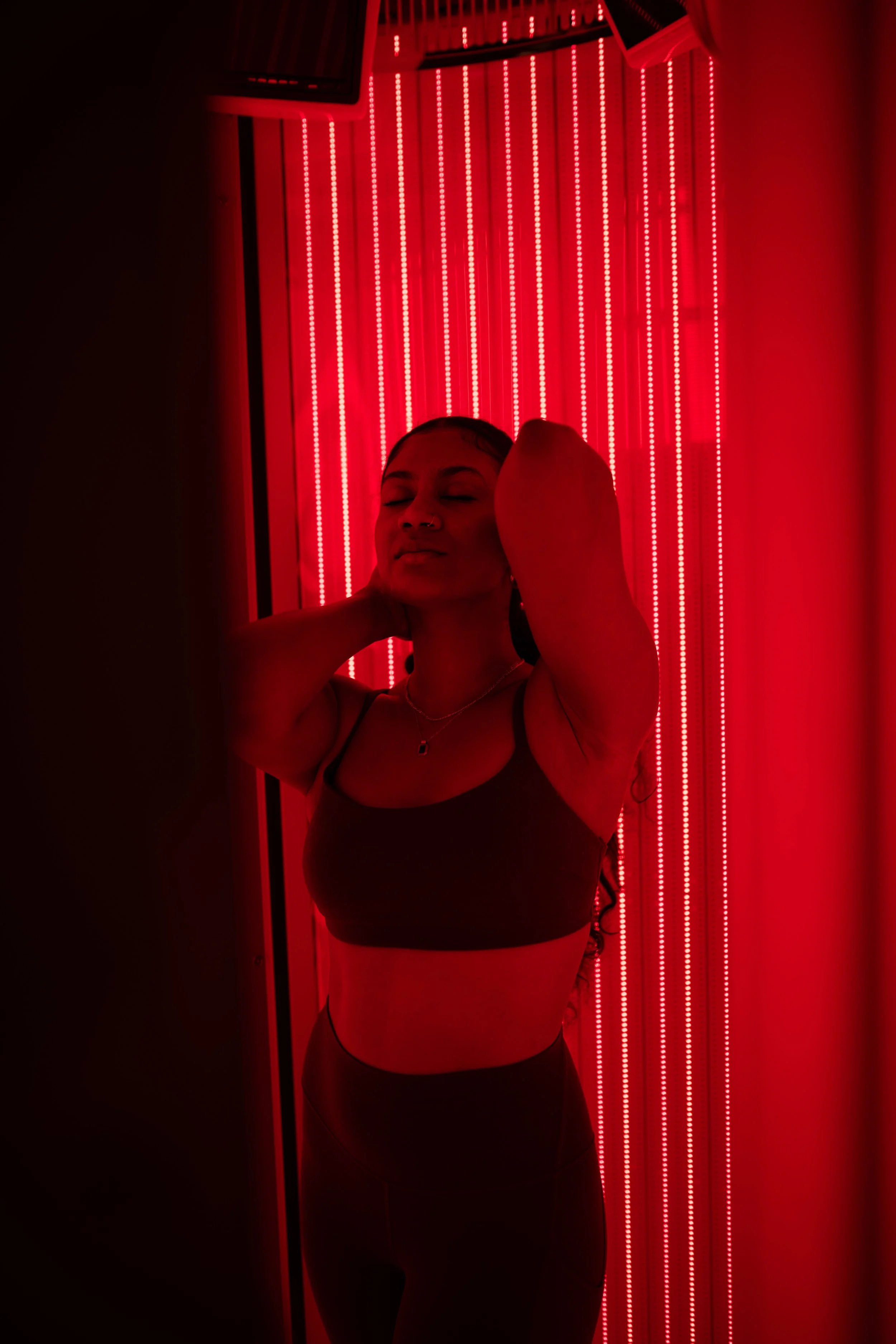 A woman standing with her eyes closed, hands behind her head, illuminated by red lights inside the red light therapy booth.