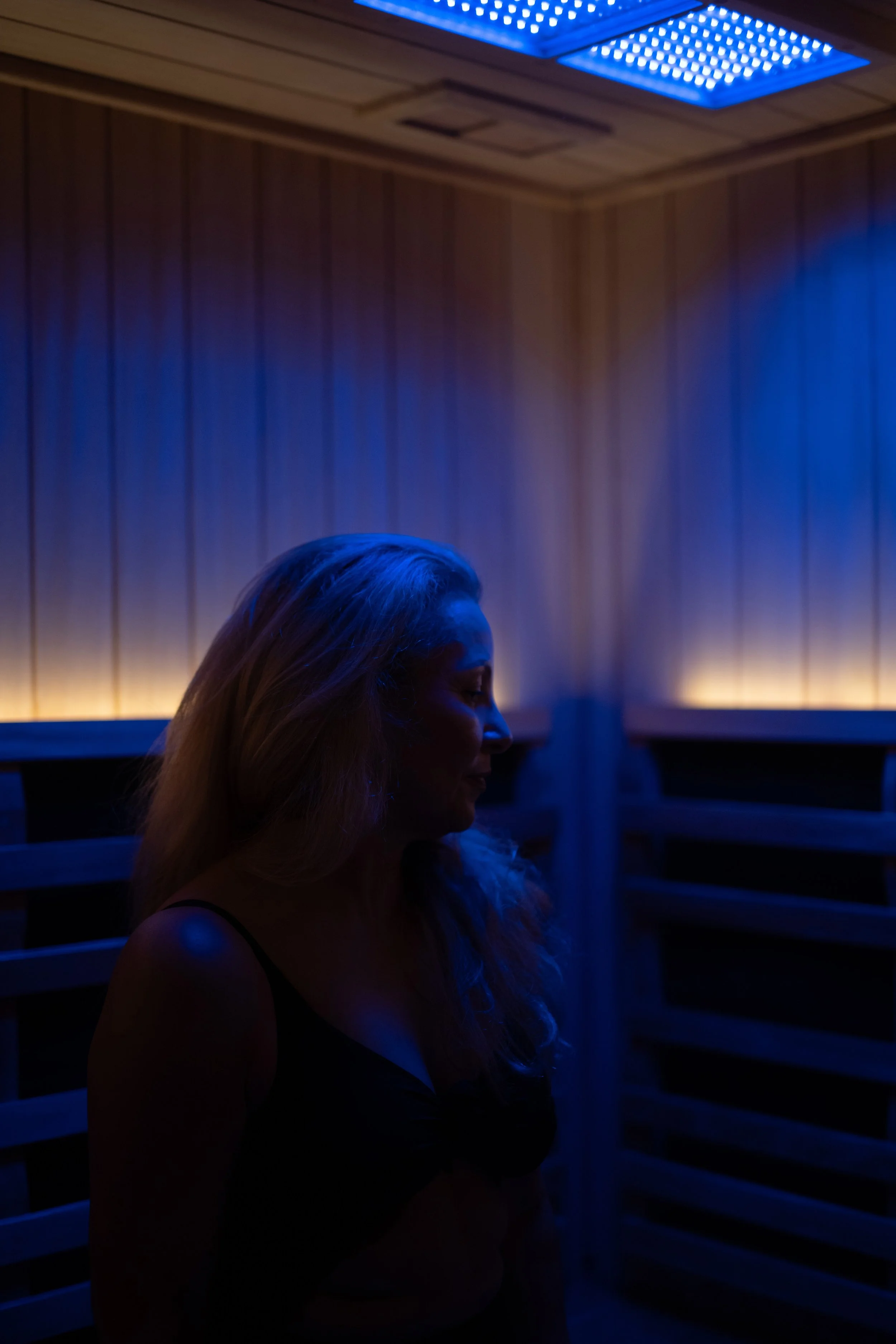 A woman with long hair in a dark sauna illuminated by blue lighting, standing near wooden slats on the wall.