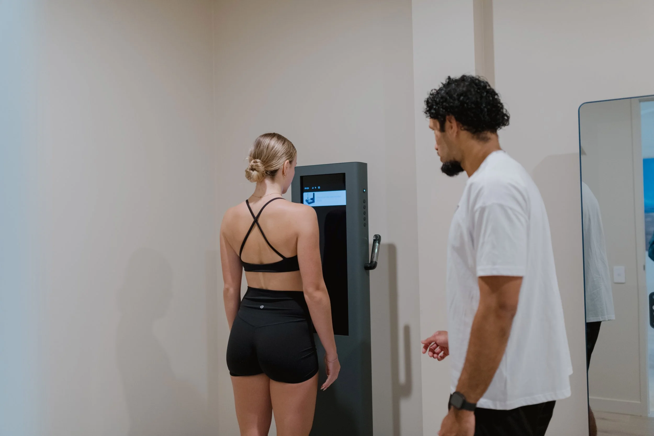 A woman with blonde hair in a bun wearing a black sports bra and shorts standing on a Visbody body composition scale, with a male health professional observing the 3D scan.