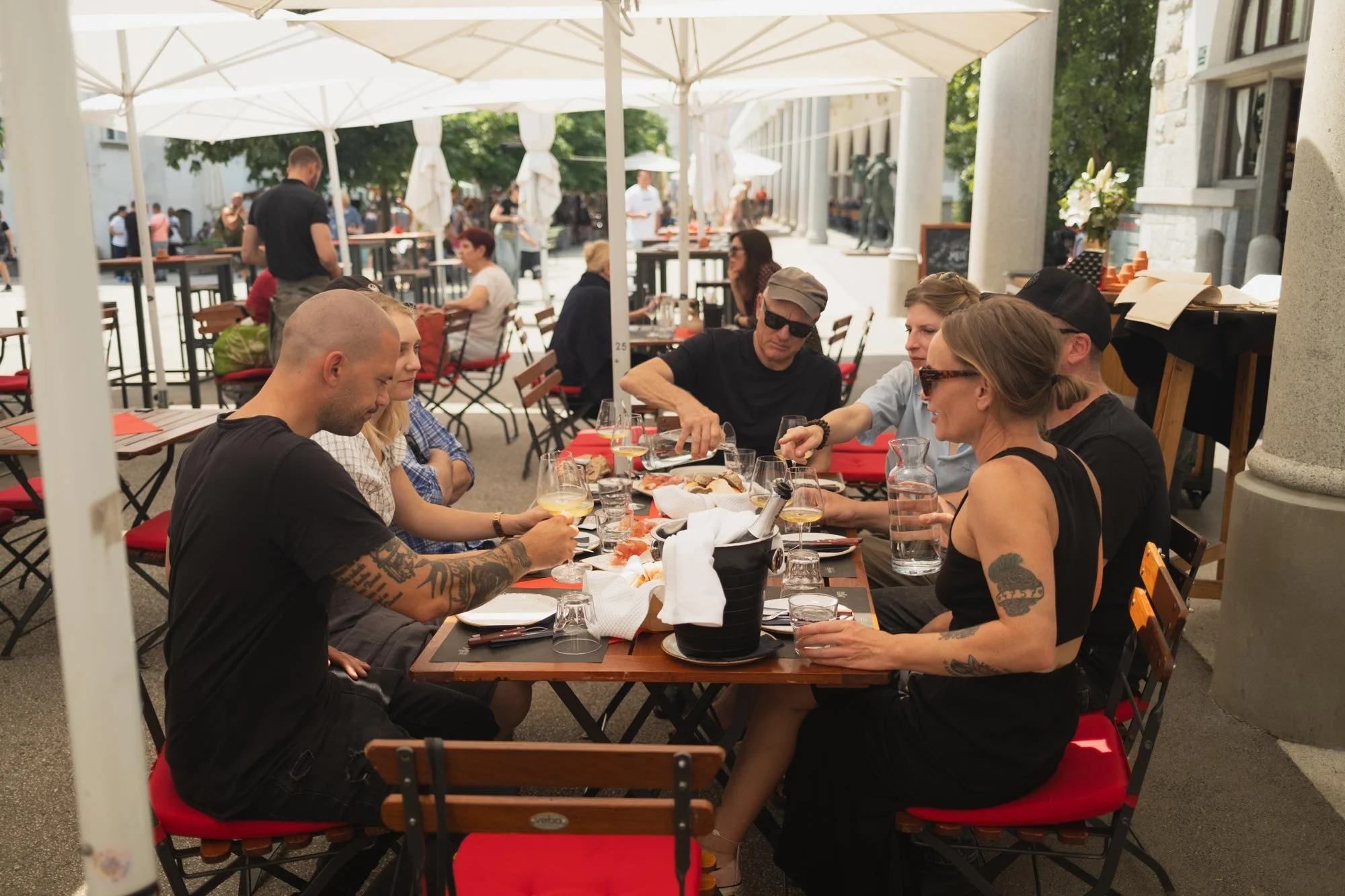 People sitting at an outdoor restaurant table enjoying a meal on a sunny day. There are books and various drinks, with a background of other diners and waitstaff walking along the street.