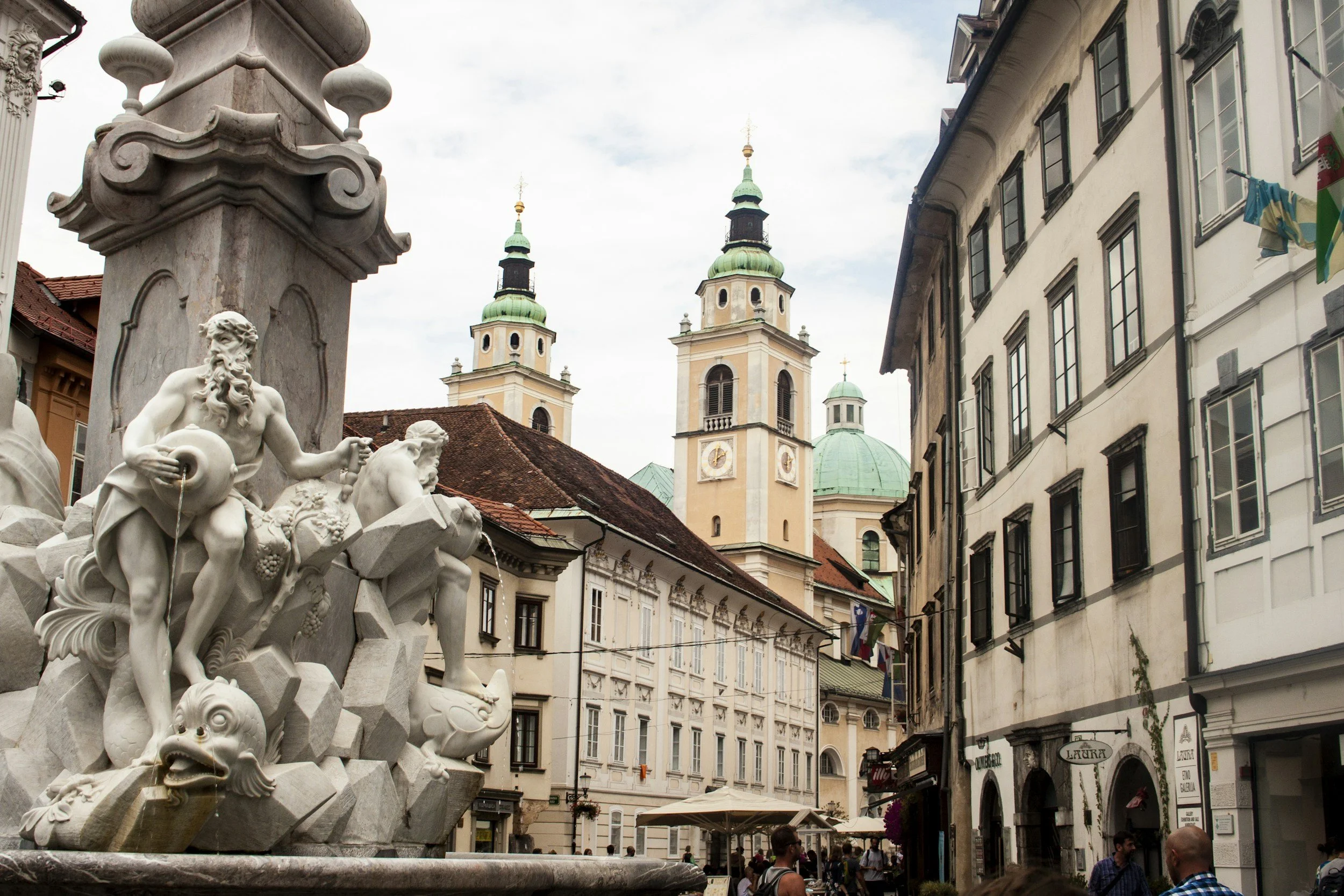 The image features a historic European town square with a detailed white marble fountain in the foreground. The fountain is decorated with mythological figures, including a bearded man and various animals, with water flowing from its mouths. In the background are two tall church towers with green domes and black spires, and surrounding the square are classic European-style buildings with white facades and multiple windows. There are also people walking and sitting in the square, and some flags hanging from the buildings.