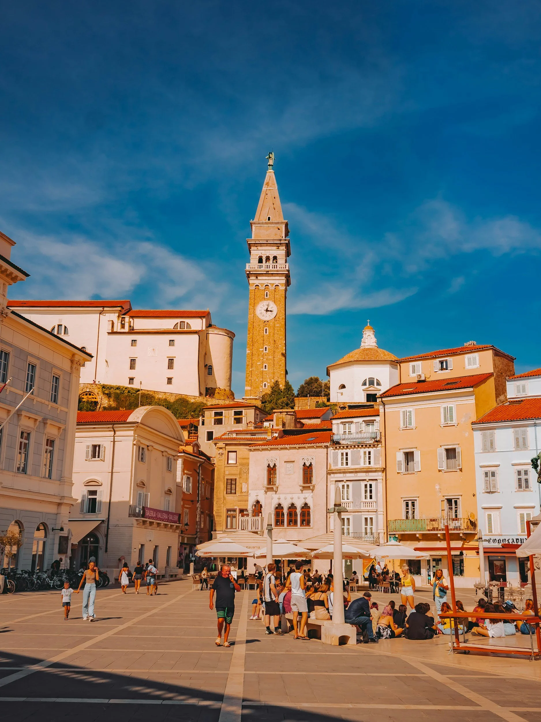 A bustling city square with people walking, sitting, and outdoor cafes beneath a tall clock tower with a statue on top, surrounded by historic buildings.