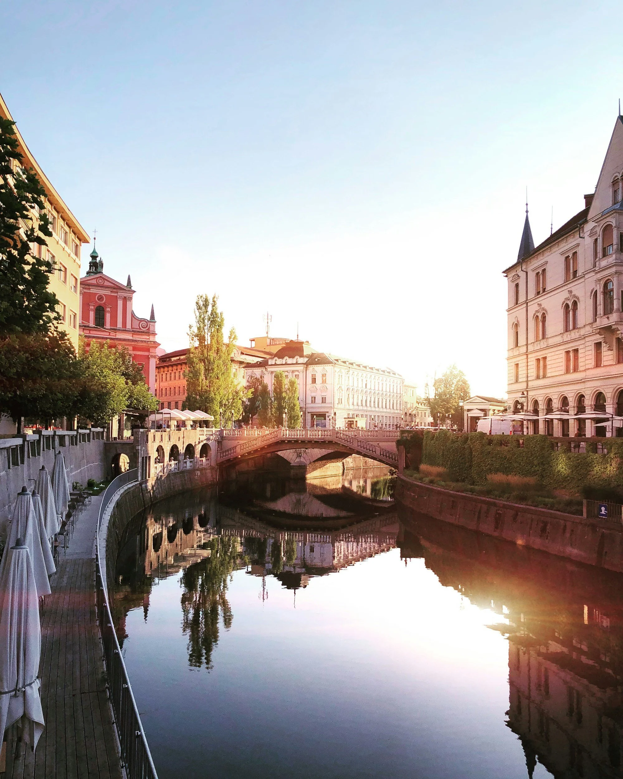 A calm city canal at sunset with reflections of historic buildings lining the water and a bridge crossing over the canal.