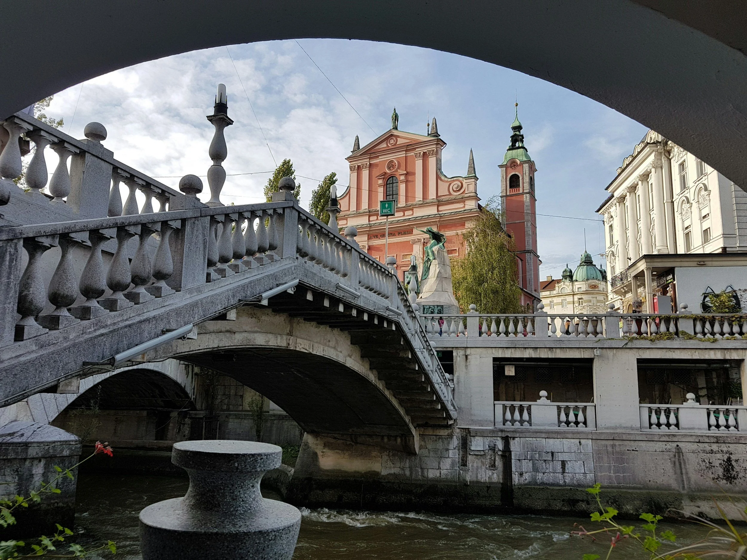 View of a historic cityscape featuring a stone bridge over a canal, with ornate buildings and churches with towers and green domes in the background. The photo is framed under a bridge arch, with a stone table and flowers visible underneath.