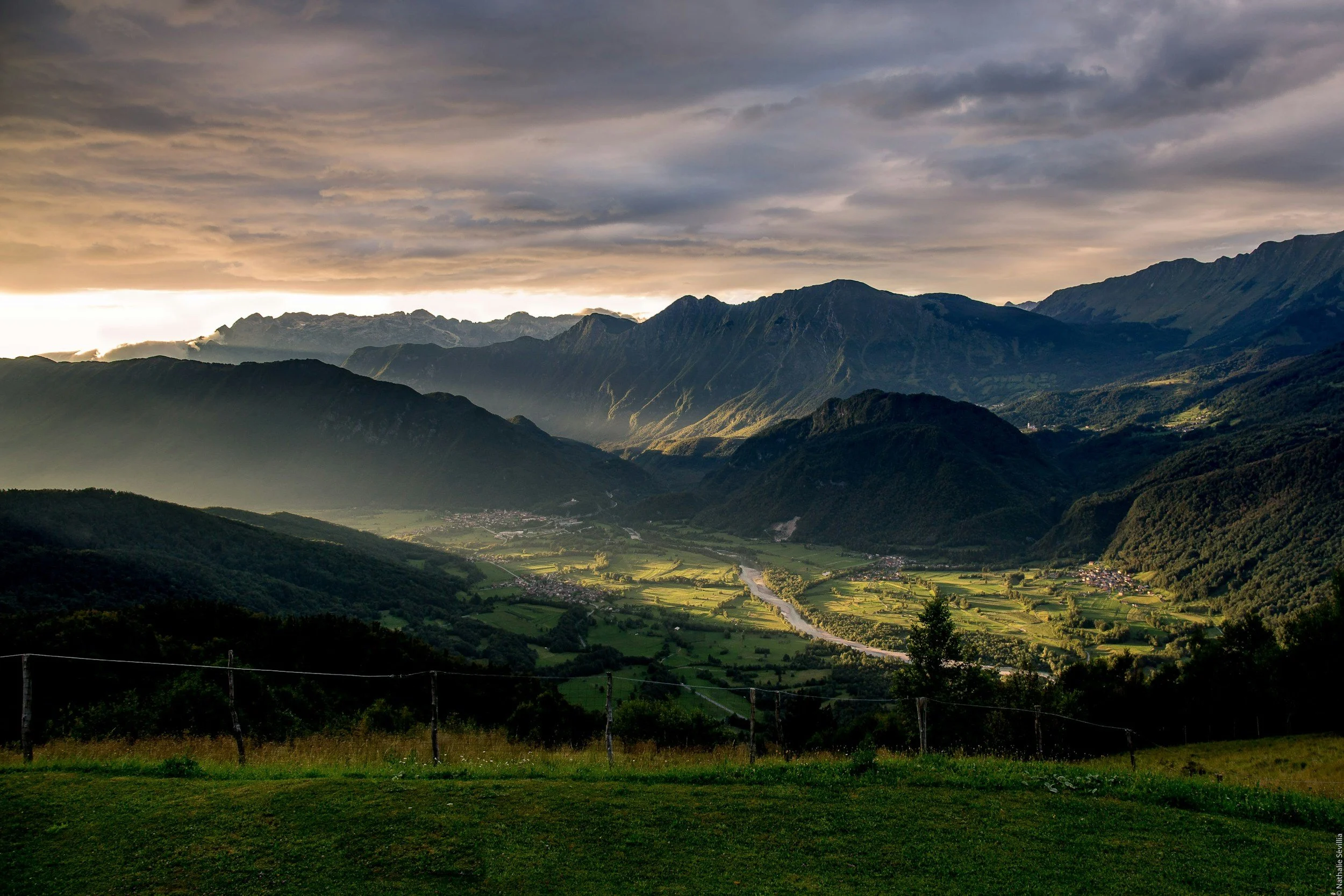 Landscape view of green valleys and rolling hills with distant mountains under a cloudy sky with evening or early morning light.