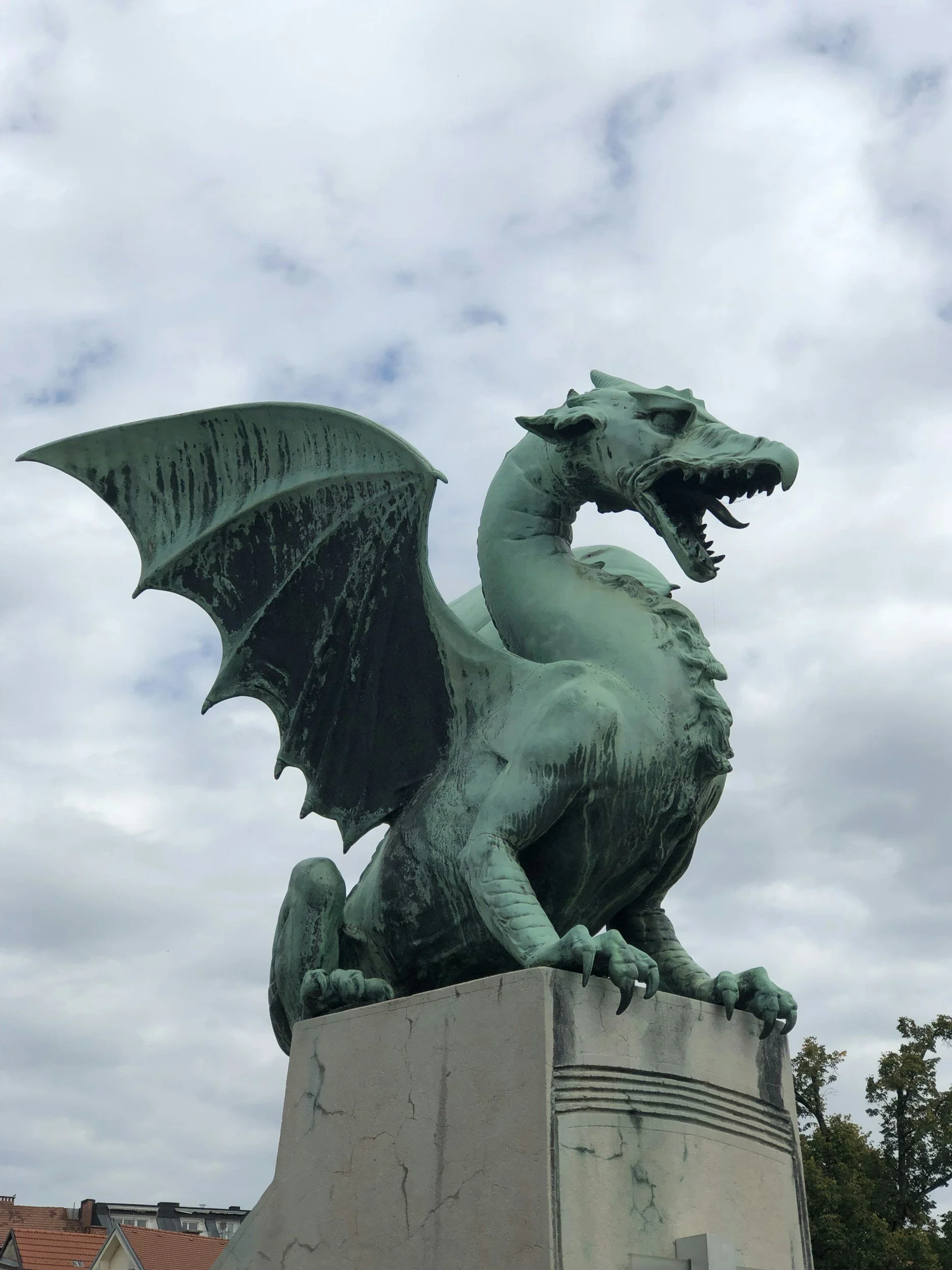 Bronze statue of a dragon with wings, perched on a pedestal against a cloudy sky.
