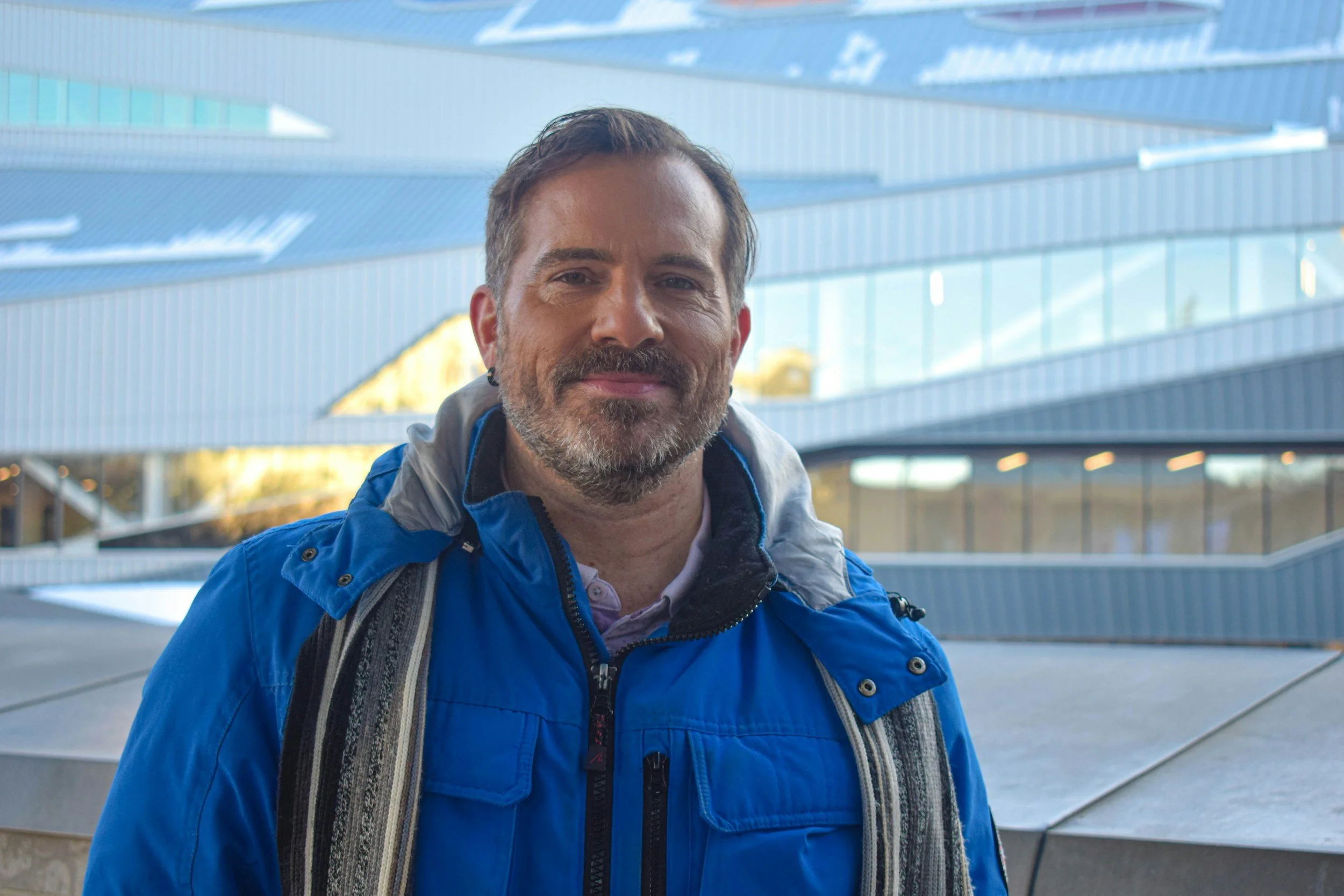 A middle-aged man with short brown hair and a beard, smiling, wearing a blue jacket and carrying a backpack, standing outdoors in front of a modern building with glass windows and metal panels.