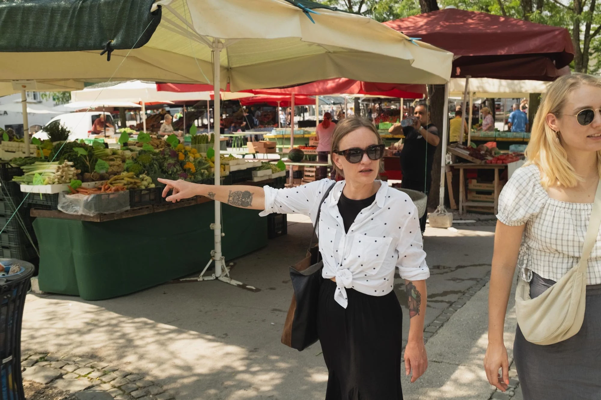 Two women are at an outdoor market with tents selling vegetables. One woman with black hair, sunglasses, and tattoos on her arm is pointing to the vegetables, while the other woman with blond hair and sunglasses is partially visible.