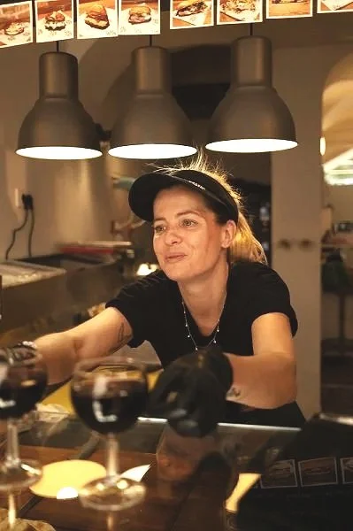 A woman working behind the counter at a restaurant or bar, with three black pendant lights overhead and menu images displayed above.
