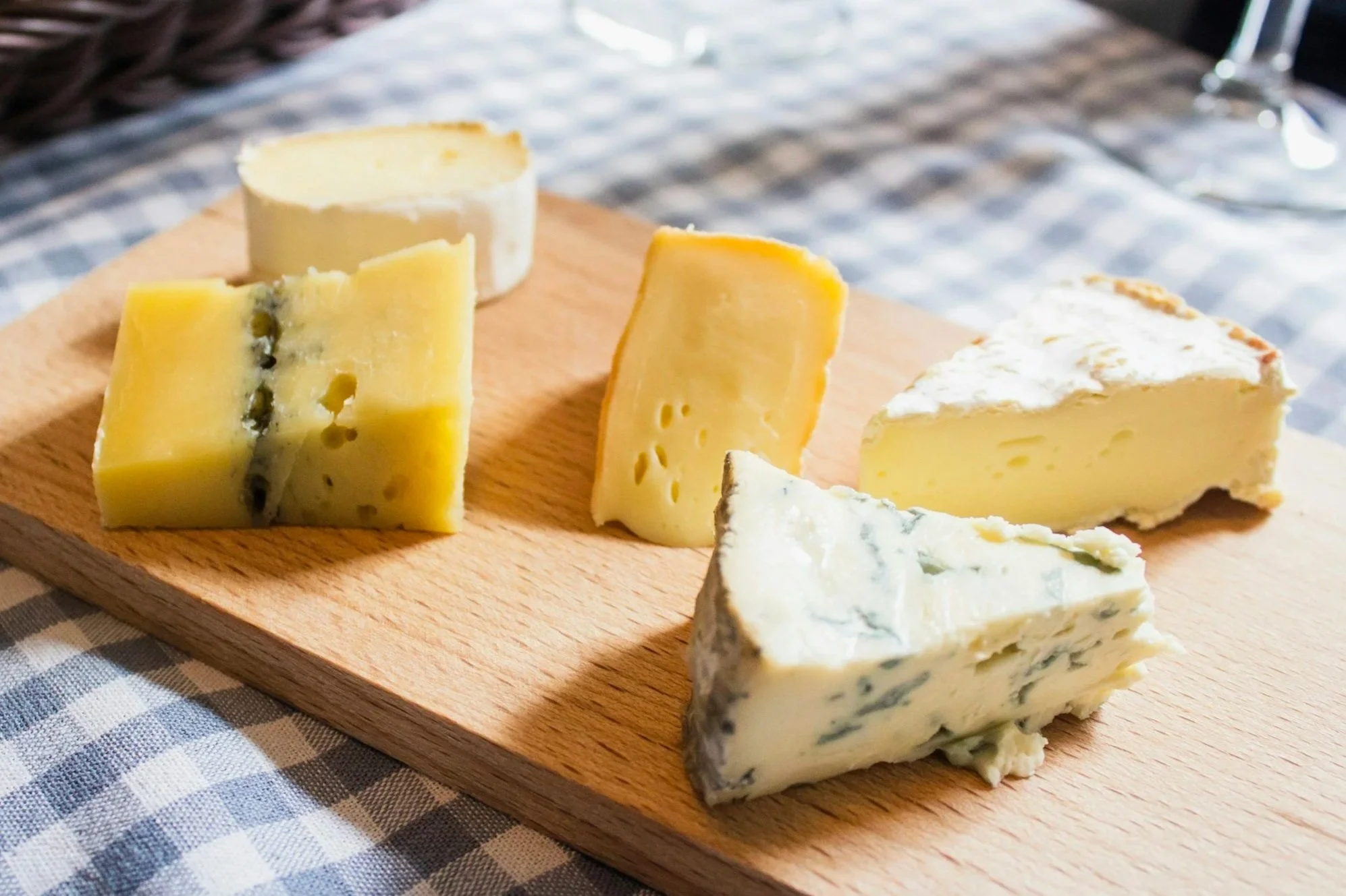 A wooden serving board with assorted cheeses, including blue cheese, cheddar, and another semi-hard cheese, placed on a checkered cloth.