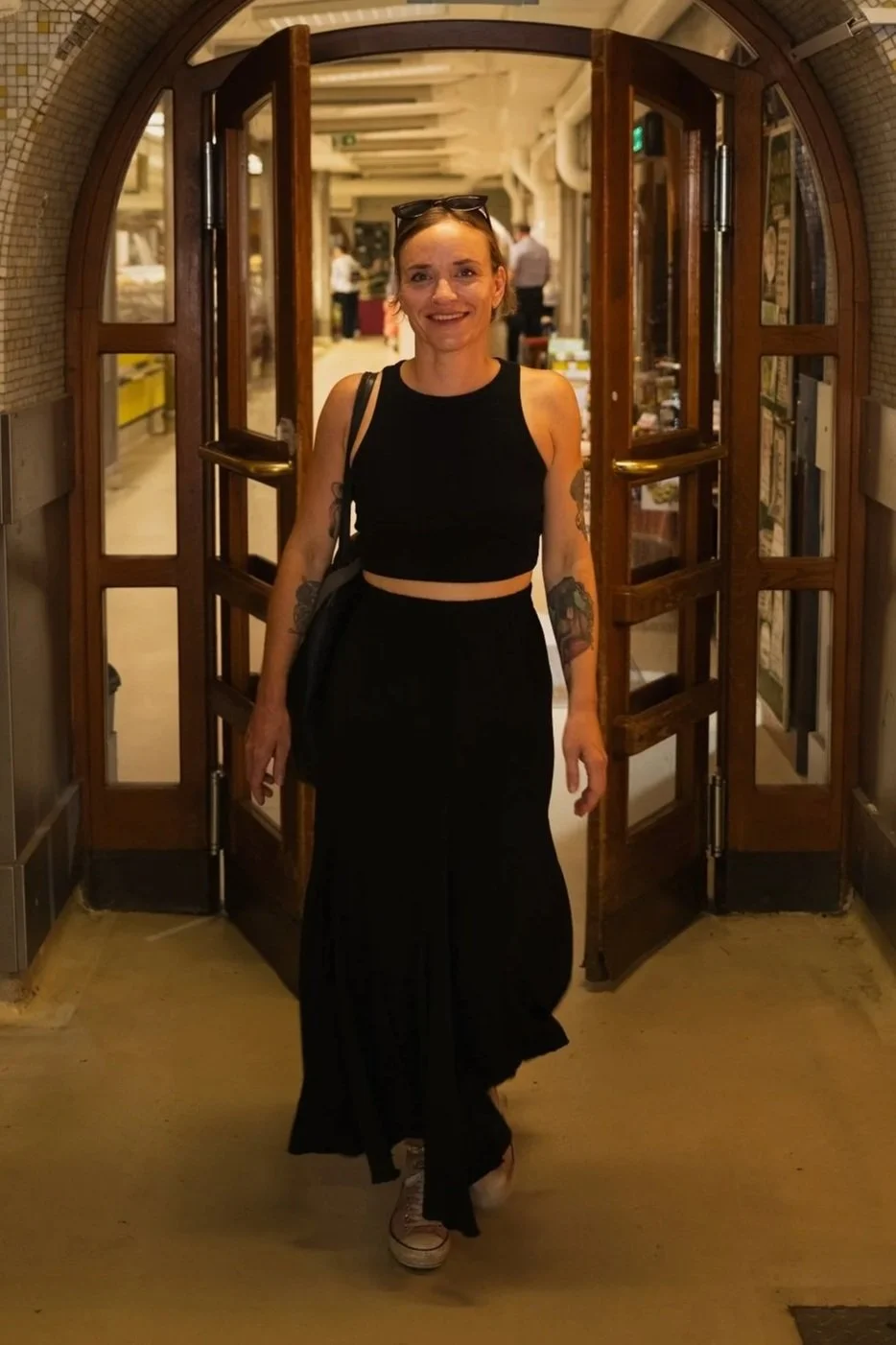 A smiling woman with tattoos on her arms, wearing a black sleeveless crop top, long black skirt, white sneakers, and sunglasses on her head, standing in front of open wooden double doors in an indoor setting filled with people and displays.