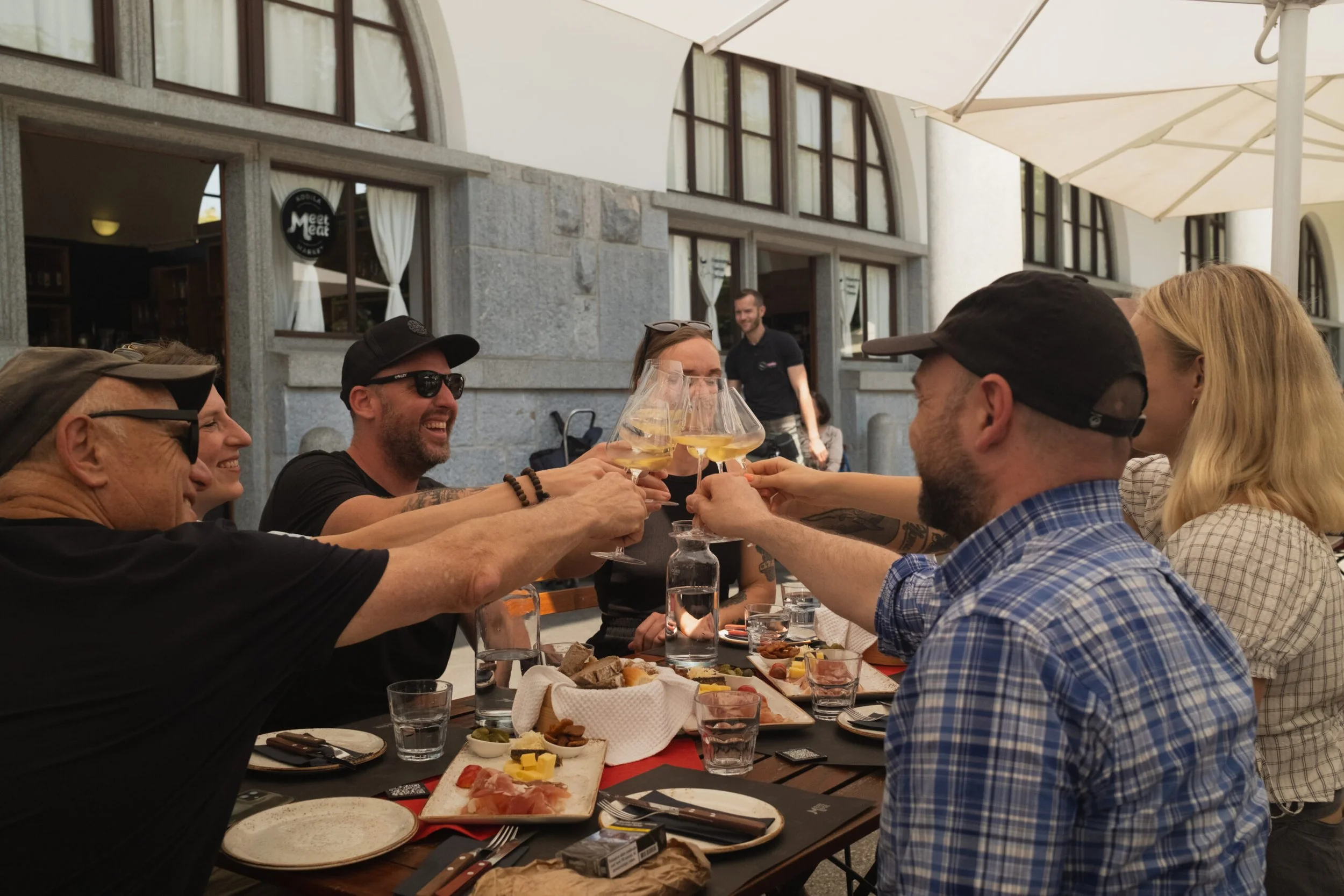 Group of people celebrating at an outdoor restaurant, raising glasses of white wine for a toast, with food and drinks on the table.