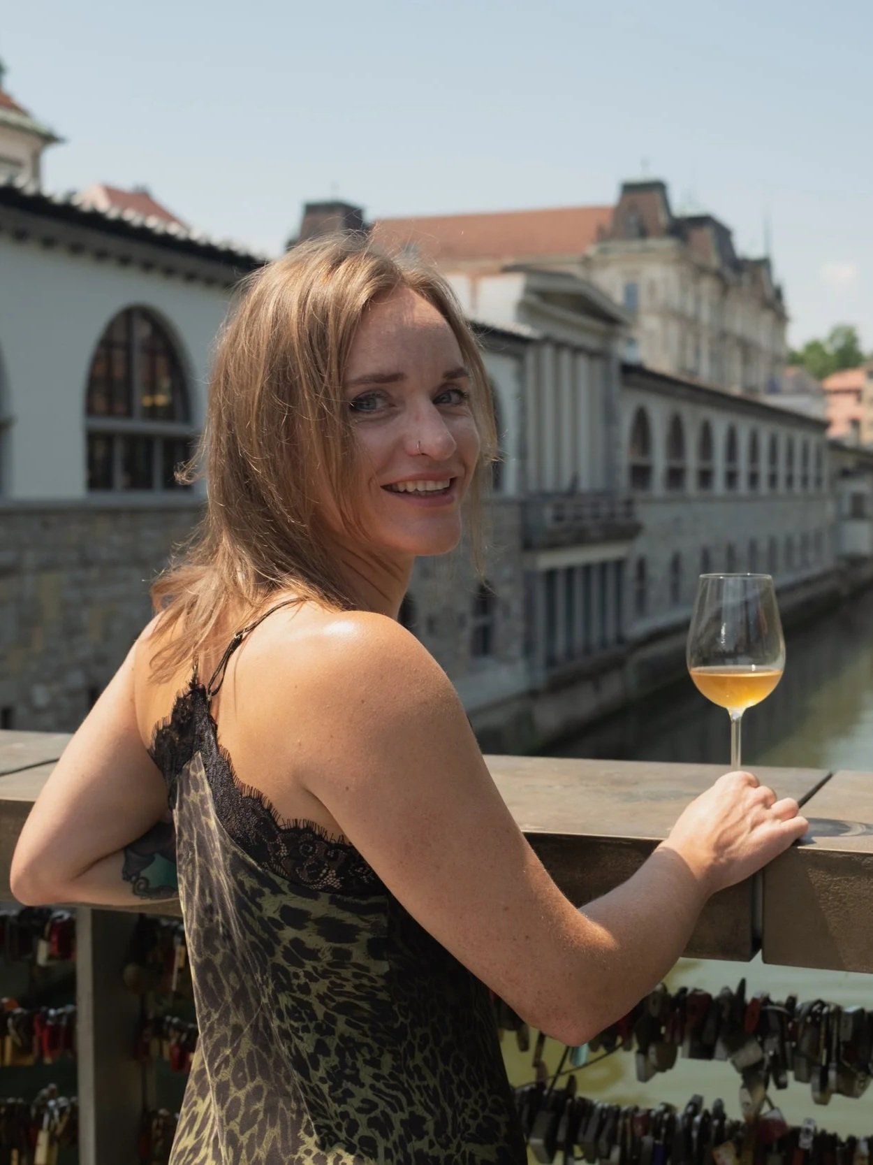A woman with shoulder-length blonde hair and a nose piercing is smiling and holding a glass of white wine on a balcony overlooking a canal with historic buildings.