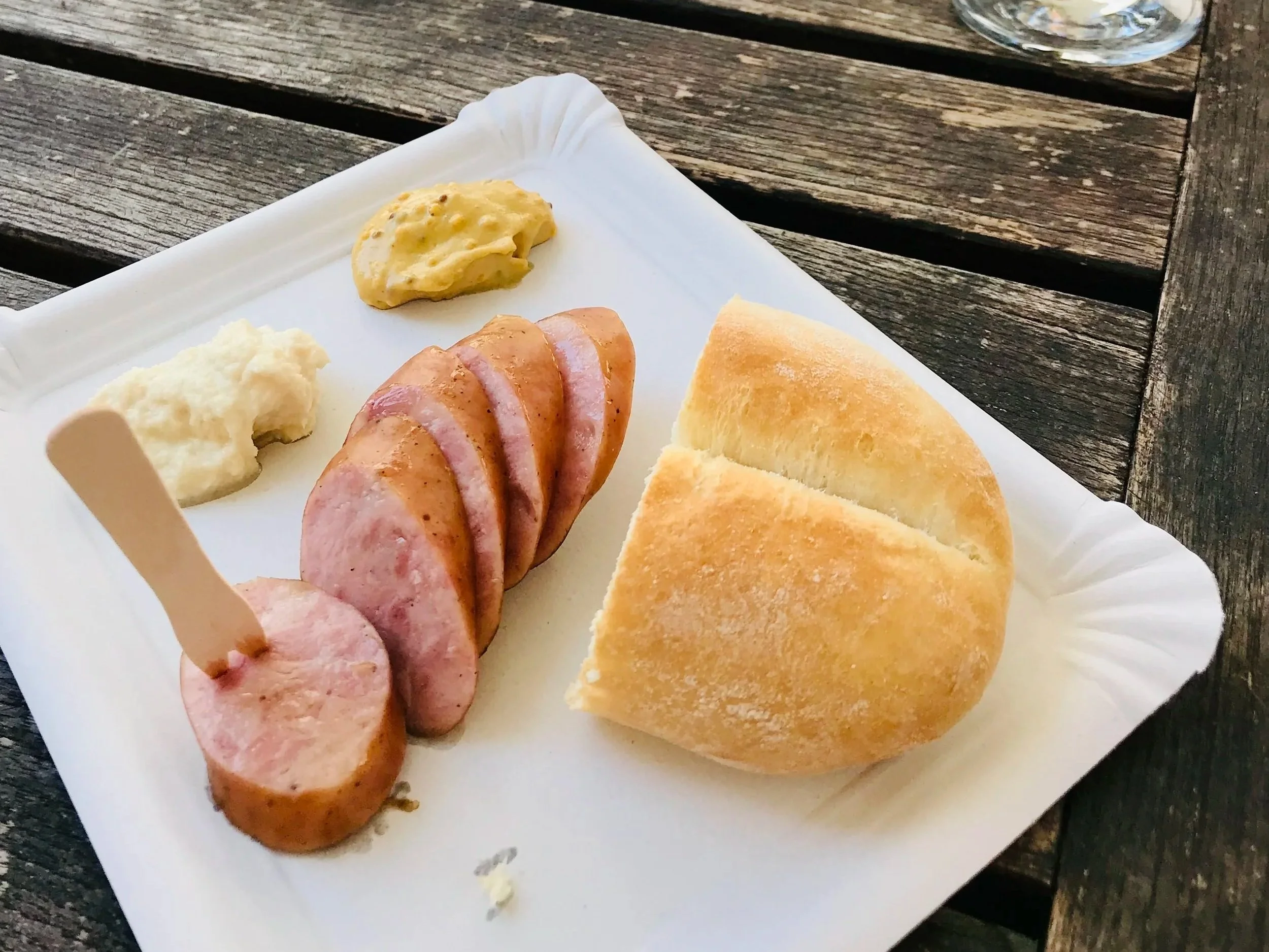 Sliced sausage, mashed potatoes, sliced bread, mustard, and a glass of water on a white paper tray on a wooden table outdoors.