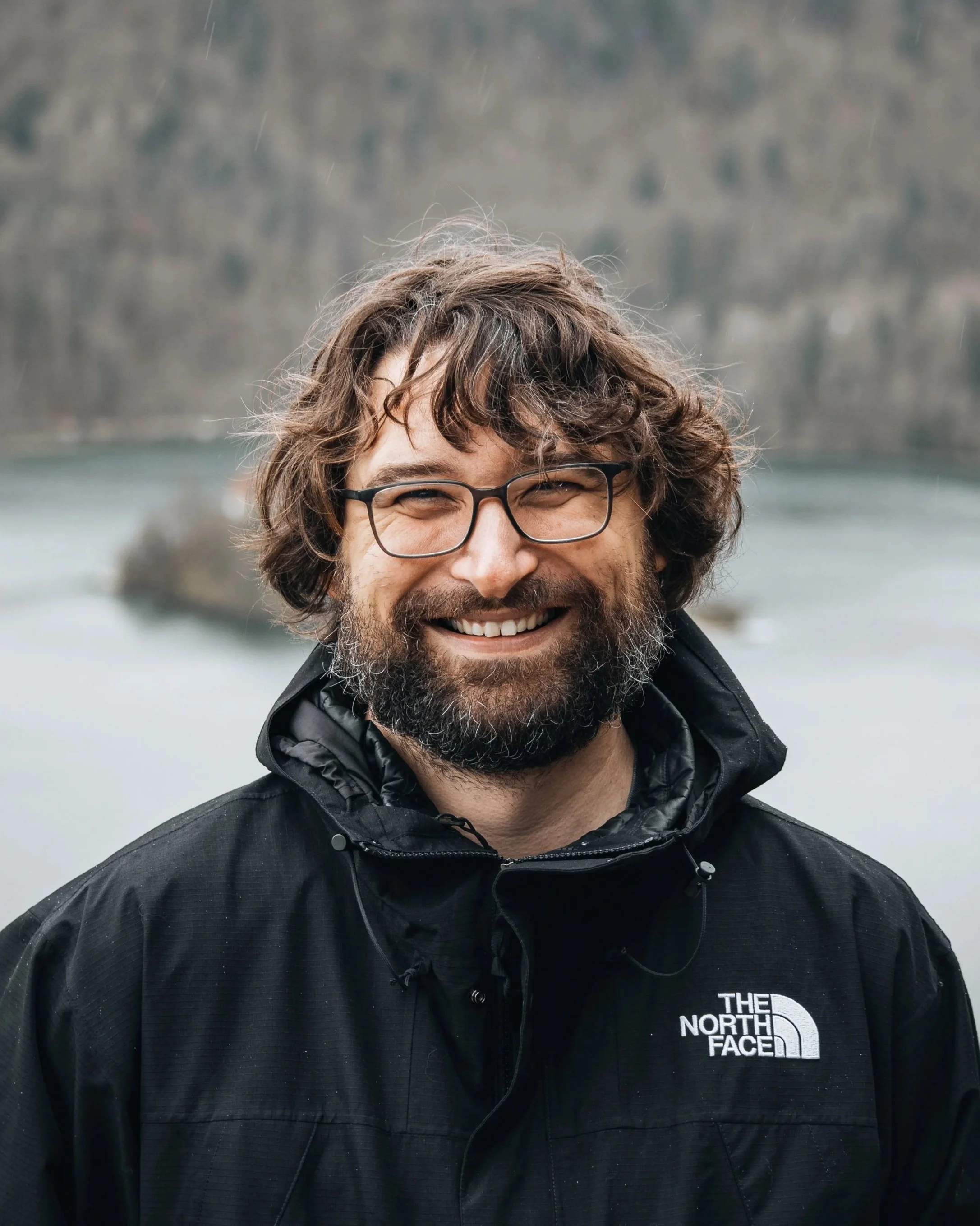 Close-up of a smiling man with glasses and curly hair, wearing a black North Face jacket, outdoors with a body of water and a forested hillside in the background.