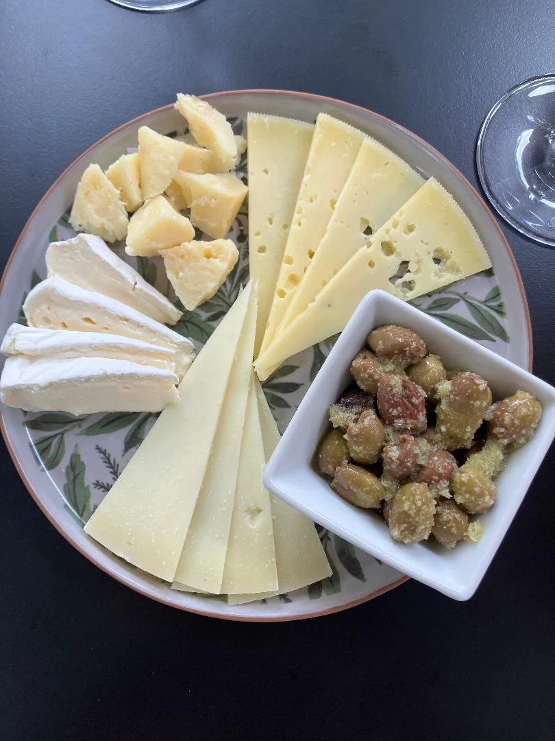 A cheese platter with various cheeses, a bowl of candied nuts, and two empty wine glasses on a black surface.