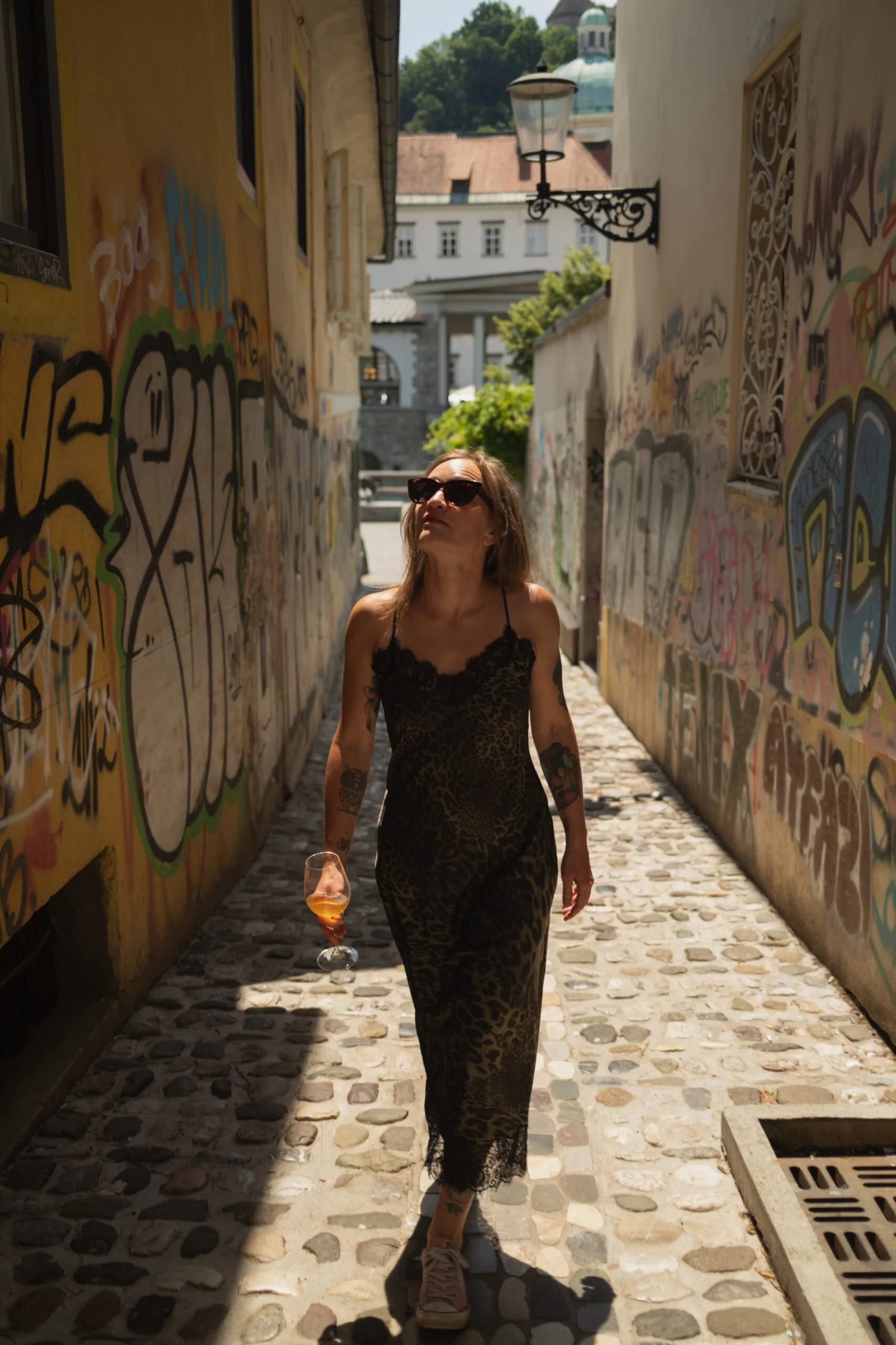 A woman with tattoos wearing a lace dress and sunglasses walking down a cobblestone alleyway, holding a glass of orange-colored beverage. Graffiti covers the walls on both sides, and there is a street lamp and buildings in the background.