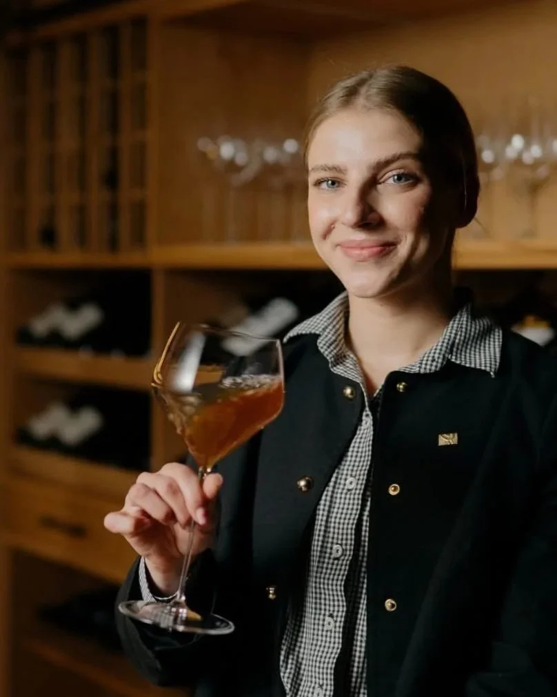 A woman with blonde hair and blue eyes smiling, holding a glass of wine in a dimly lit wine cellar or bar with shelves of wine bottles in the background.