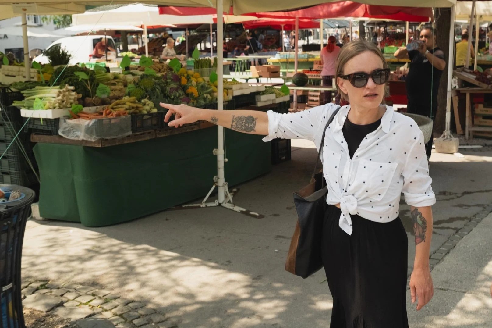 A woman wearing black sunglasses, a white shirt with black polka dots tied in a knot, and black pants is pointing towards a fruit and vegetable stand at an outdoor market. She has tattoos on her arm and is carrying a black shoulder bag.