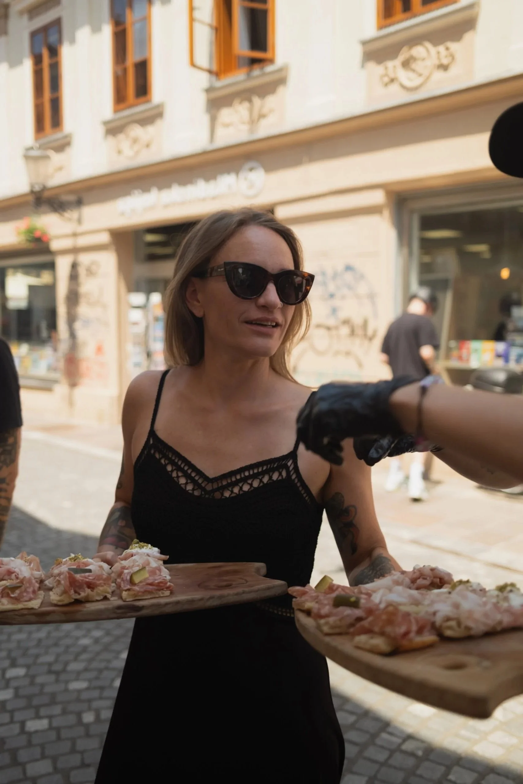 A woman wearing large sunglasses and a black crochet dress is serving food at an outdoor event, with pizza on a wooden tray.