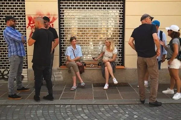 People sitting and standing outside in front of a building with graffiti and a window.