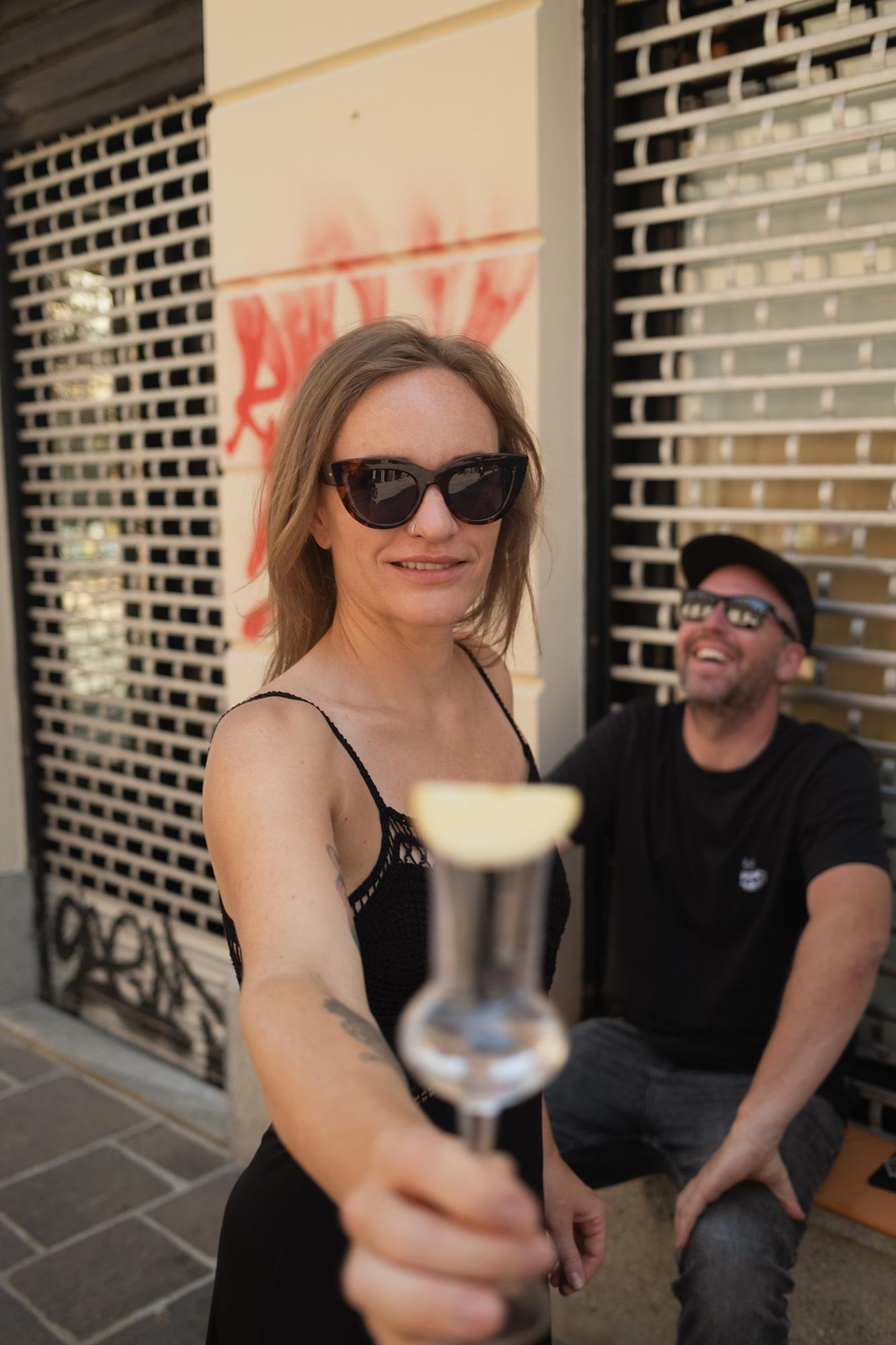 A woman wearing sunglasses and a black dress holding a drink in focus, with a man in the background sitting outdoors near a graffiti-covered wall and a black metal grid, smiling and wearing sunglasses and a cap.