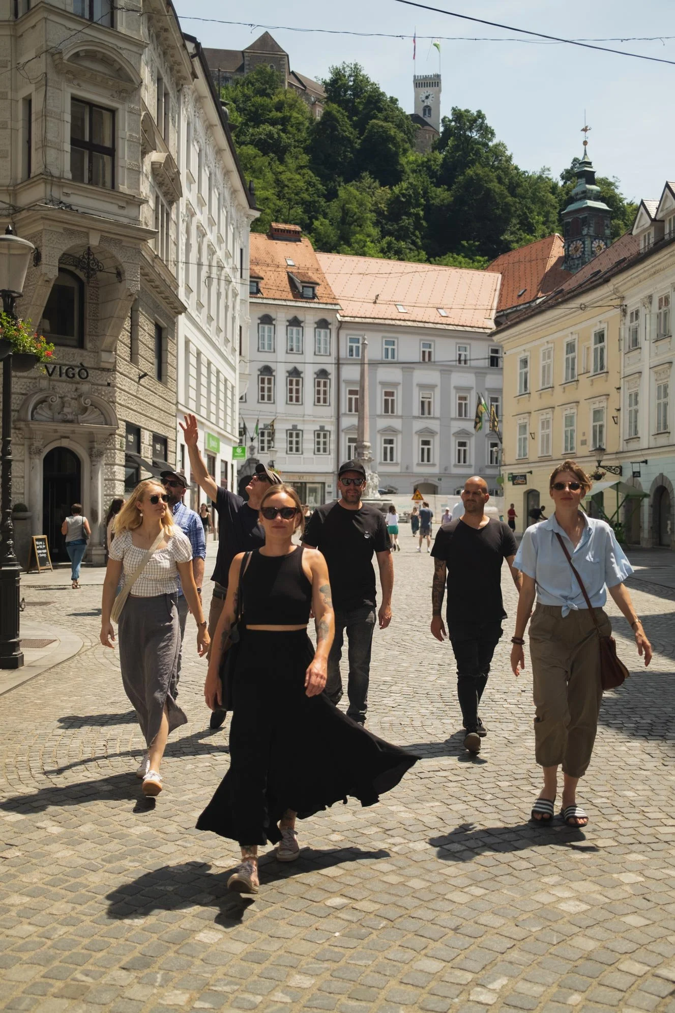 A group of people walking on a cobblestone street in a European city with historic buildings, a church tower, and a castle in the background.