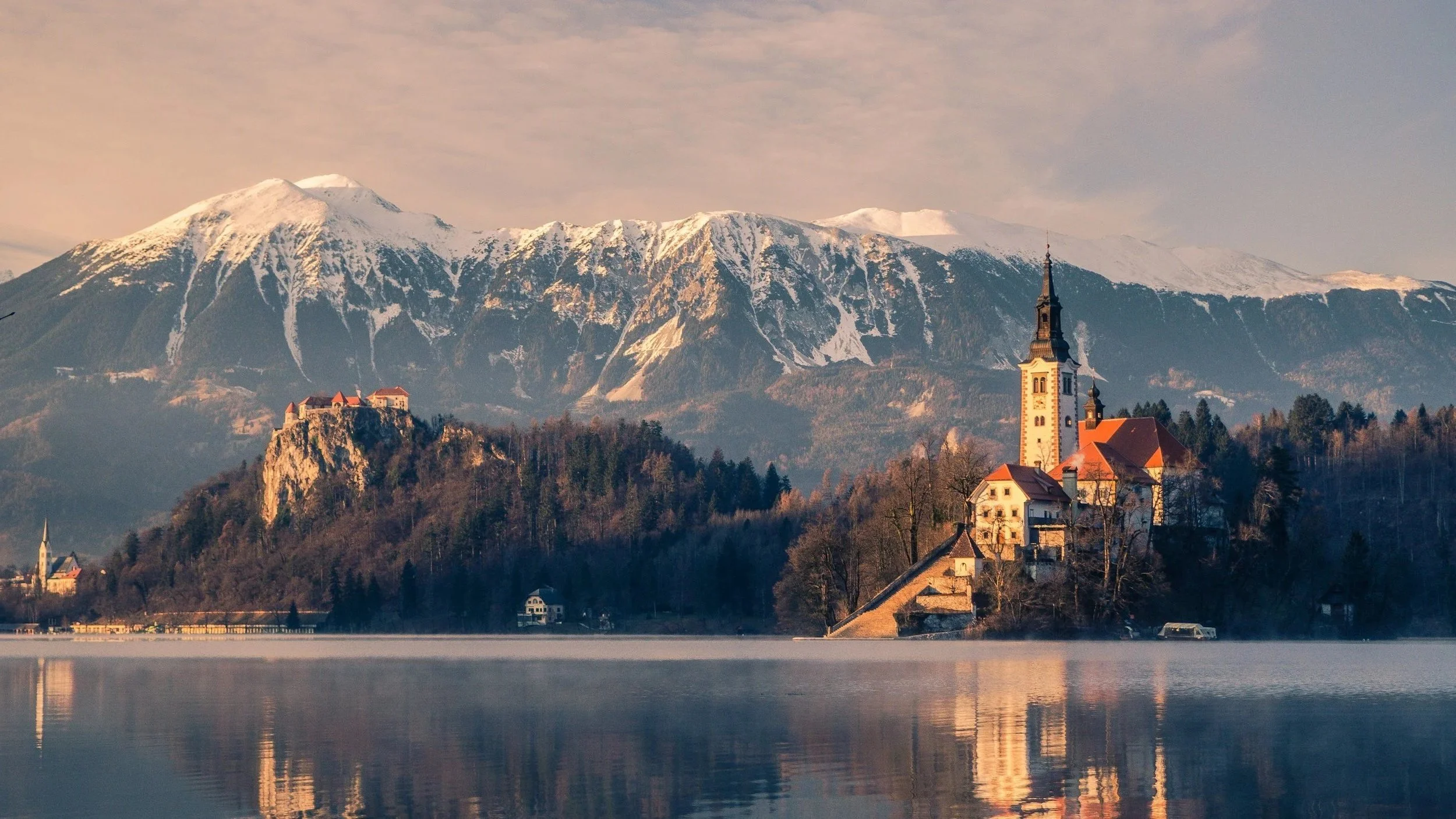 A scenic view of a lake with a small town, including a prominent church with a tall steeple, set against a backdrop of snow-capped mountains during sunset.