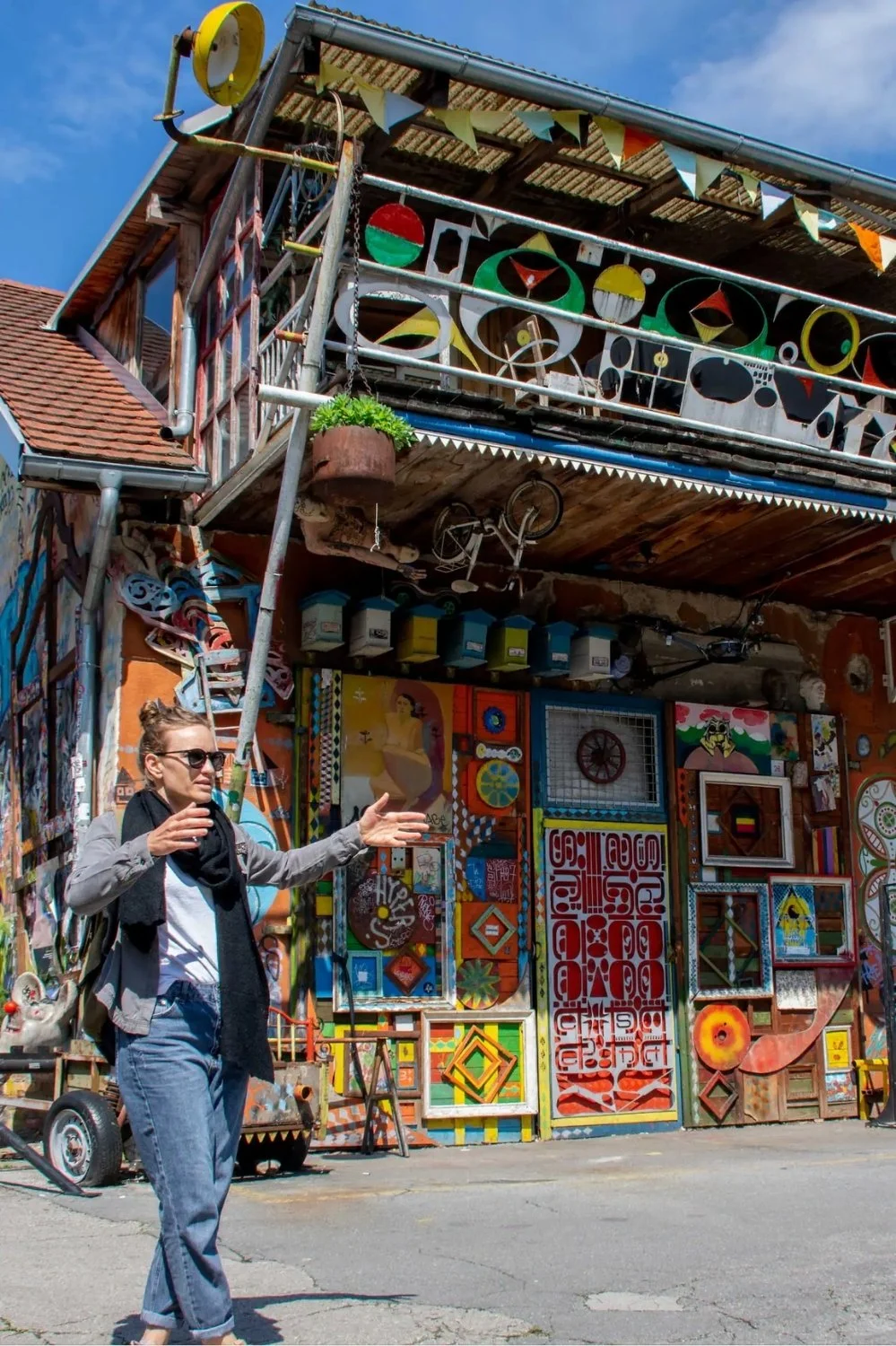 Woman with sunglasses and gray jacket gesturing in front of a colorful, decorated wall with various artwork and painting, outdoors on a sunny day.