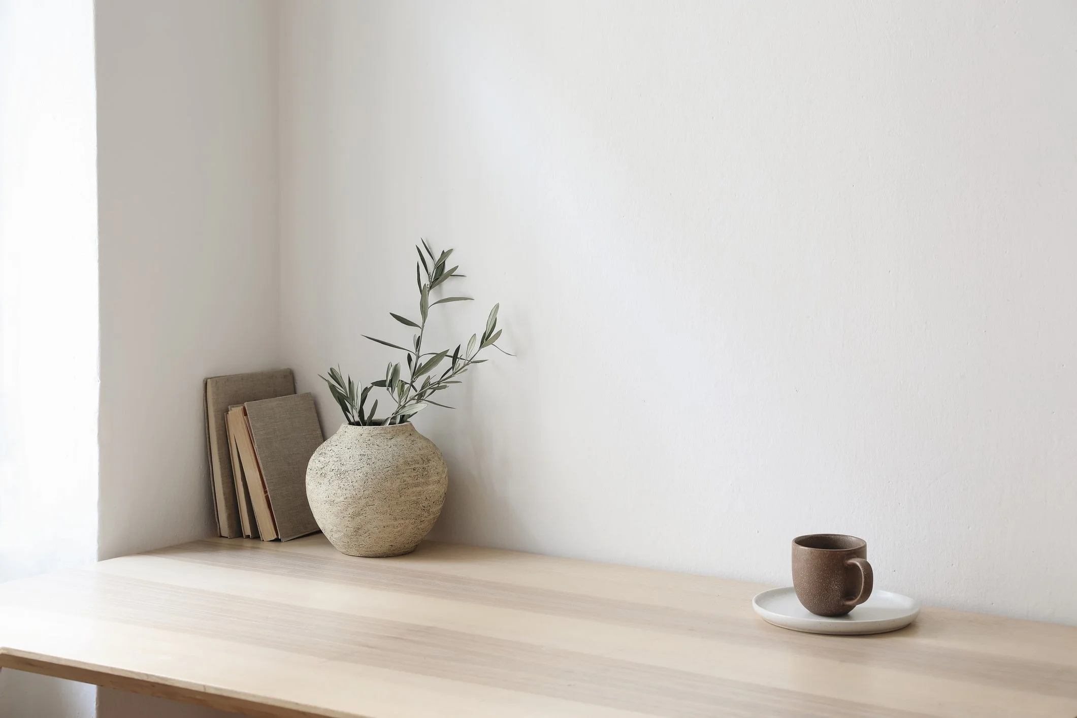 A minimalist interior with a light wooden table, a beige textured ceramic vase with a green leafy plant, a few books, a dark brown ceramic mug on a white saucer, and a white wall background.