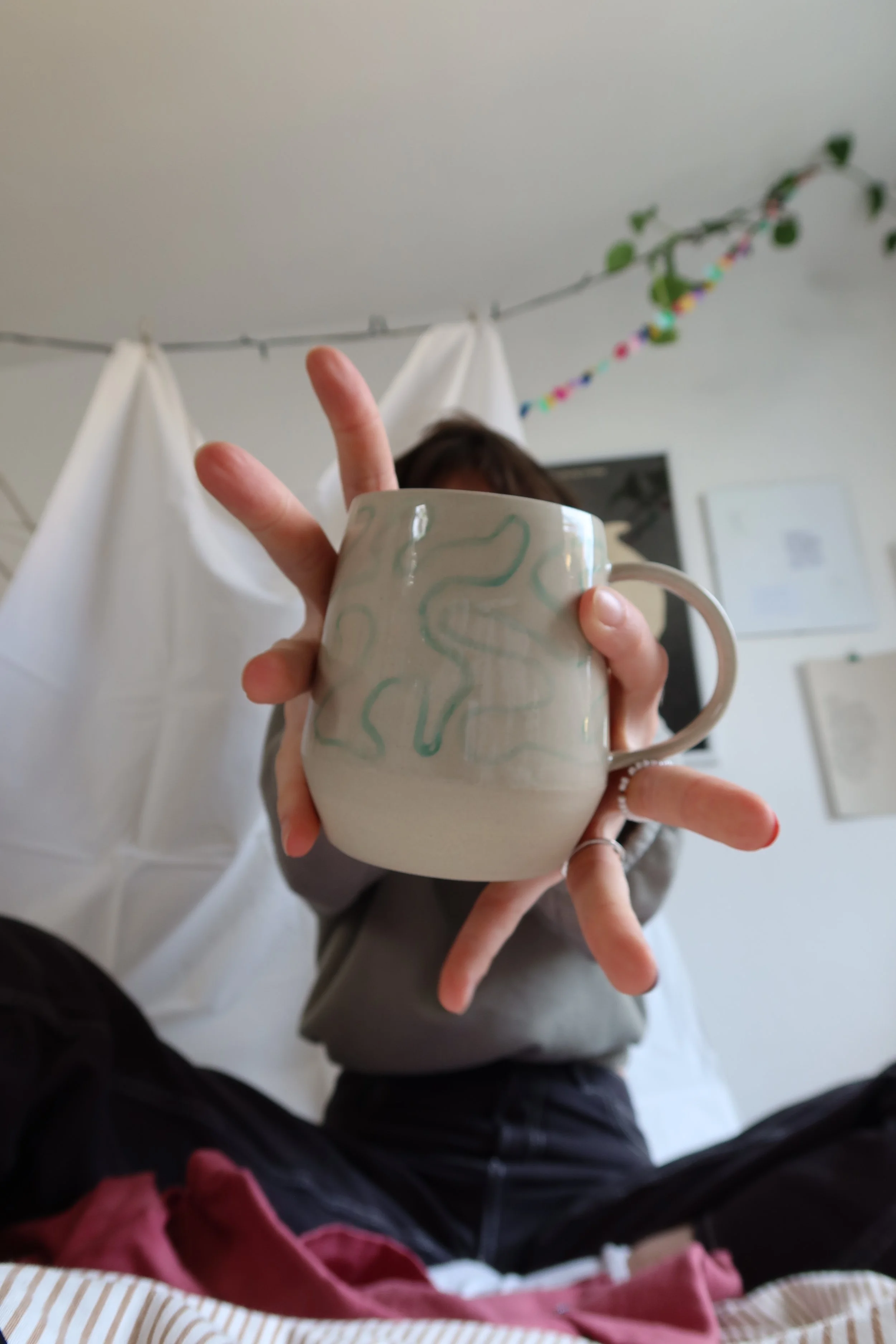 Person hält eine weiße Tasse mit zartem, grünem Muster, versteckt ihr Gesicht hinter der Tasse, im Hintergrund sind weiße Vorhänge und eine Wand mit Bildern und einer bunten Girlande.