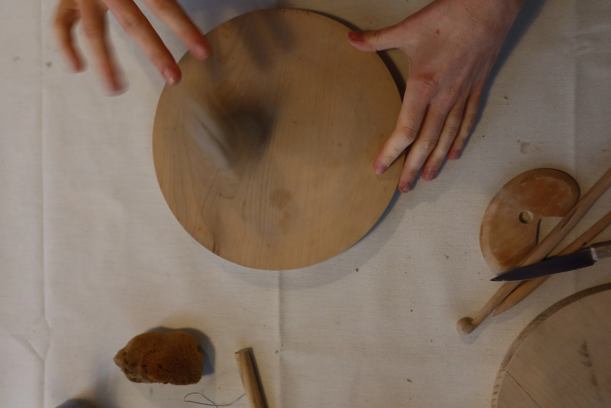 Personen formen mit den Händen Ton auf einer runde Holzscheibe auf einem Tisch, umgeben von Werkzeugen, darunter Pinsel, ein Messer und eine Töpferschlinge.
