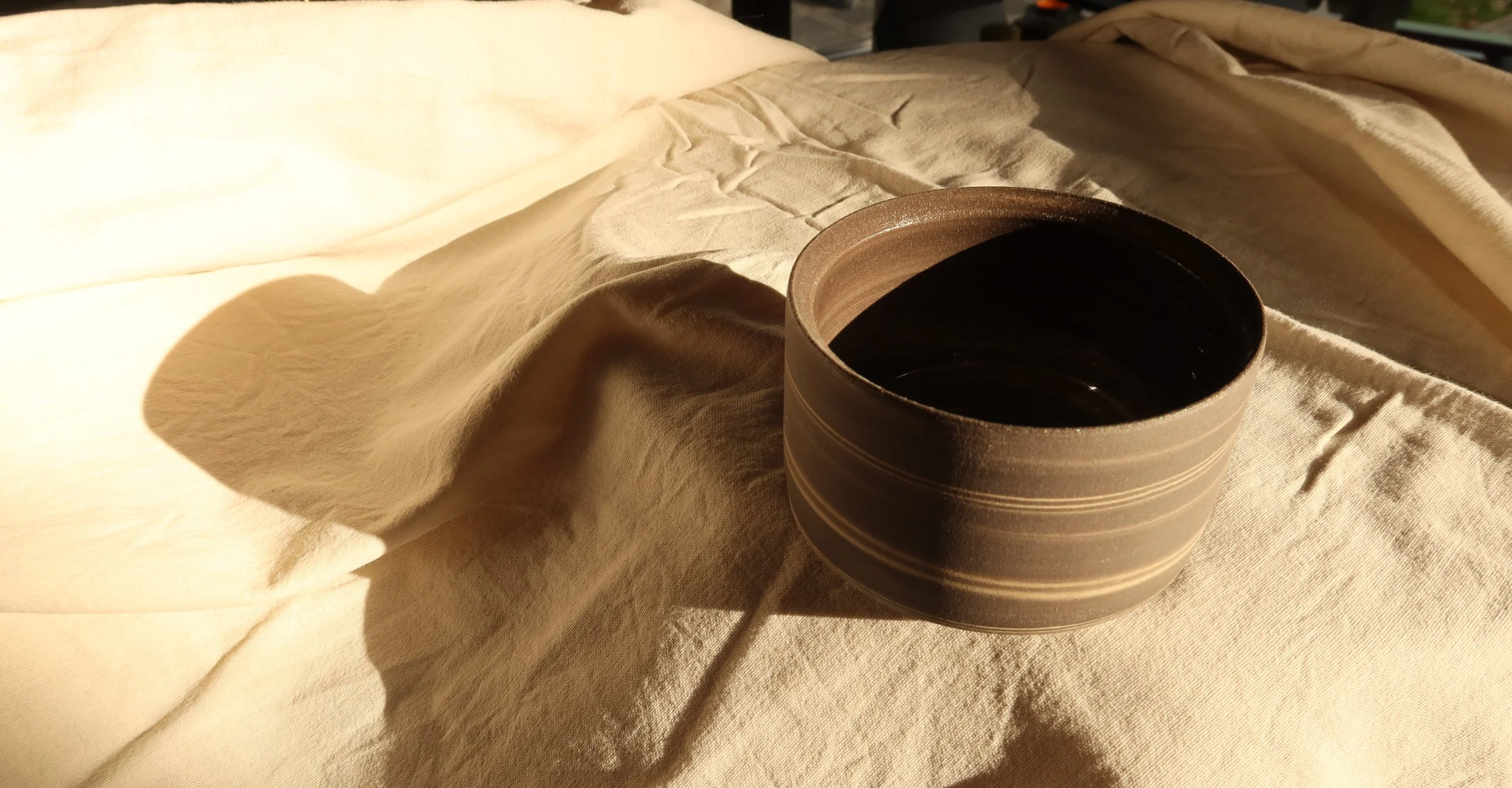 Eine schwarze Tee- oder Cappucino-Schale mit breiten, hellen Linien auf einem beige-leinenen Stoff, im Schatten und warmem Licht
